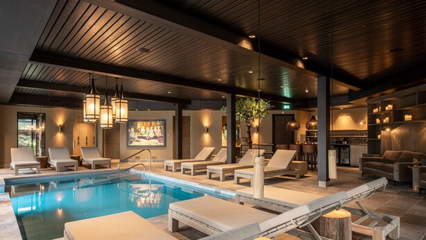 A modern indoor pool area with lounge chairs, a wooden ceiling, and ambient lighting.
