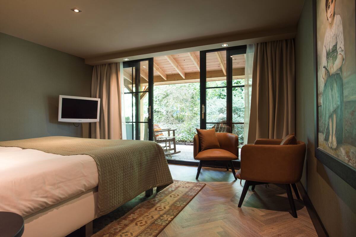 Una habitación de hotel con cama, televisor, sillón y un amplio ventanal con cortinas.