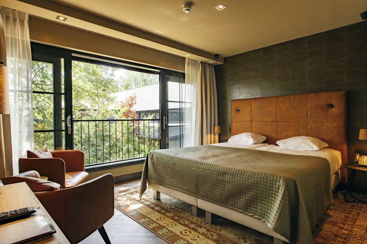Un dormitorio moderno con una cama grande, vista a zonas verdes y un rincón de descanso con mesa y sillas.