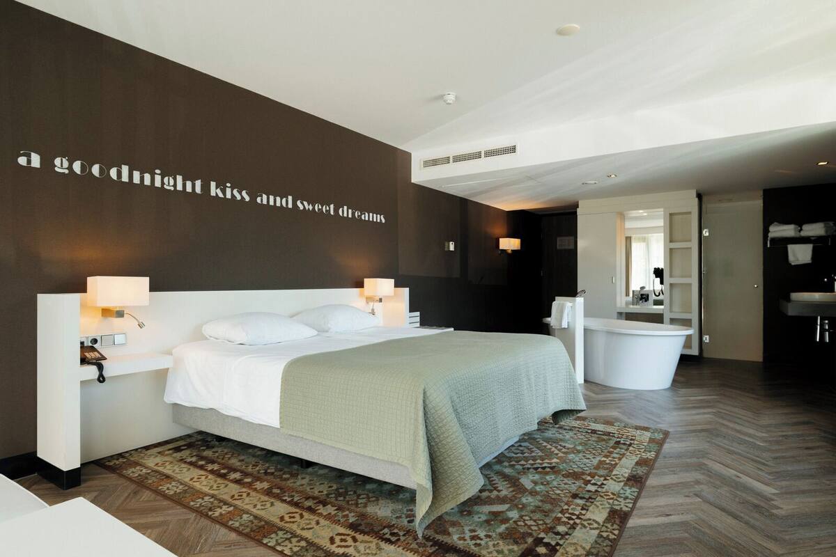 Una habitación de hotel moderna con una cama grande, lámparas de noche, un baño con bañera y una pared con una cita motivacional.