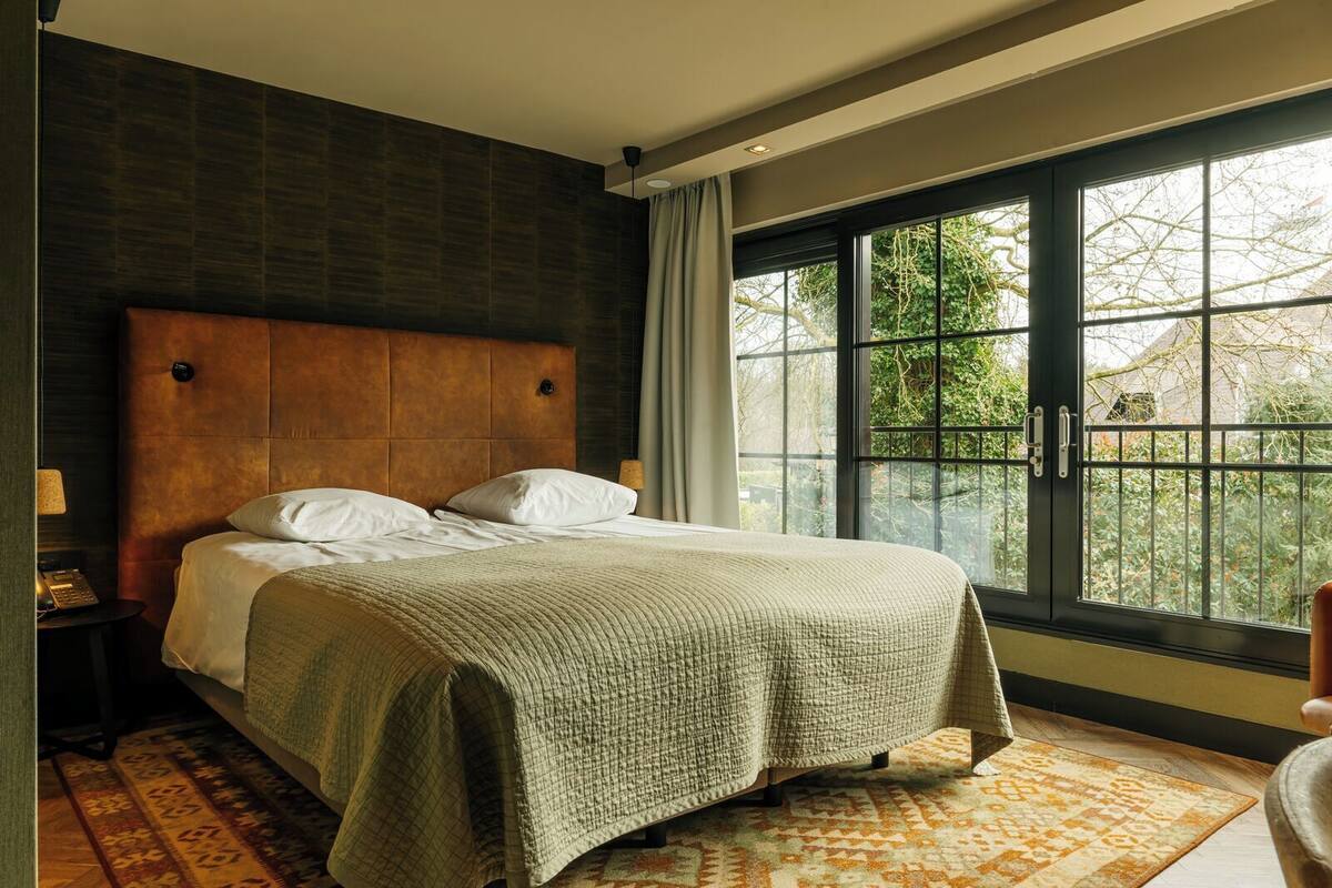 Un dormitorio con una cama grande, una mesita de noche, una lámpara y una ventana con vistas a árboles.