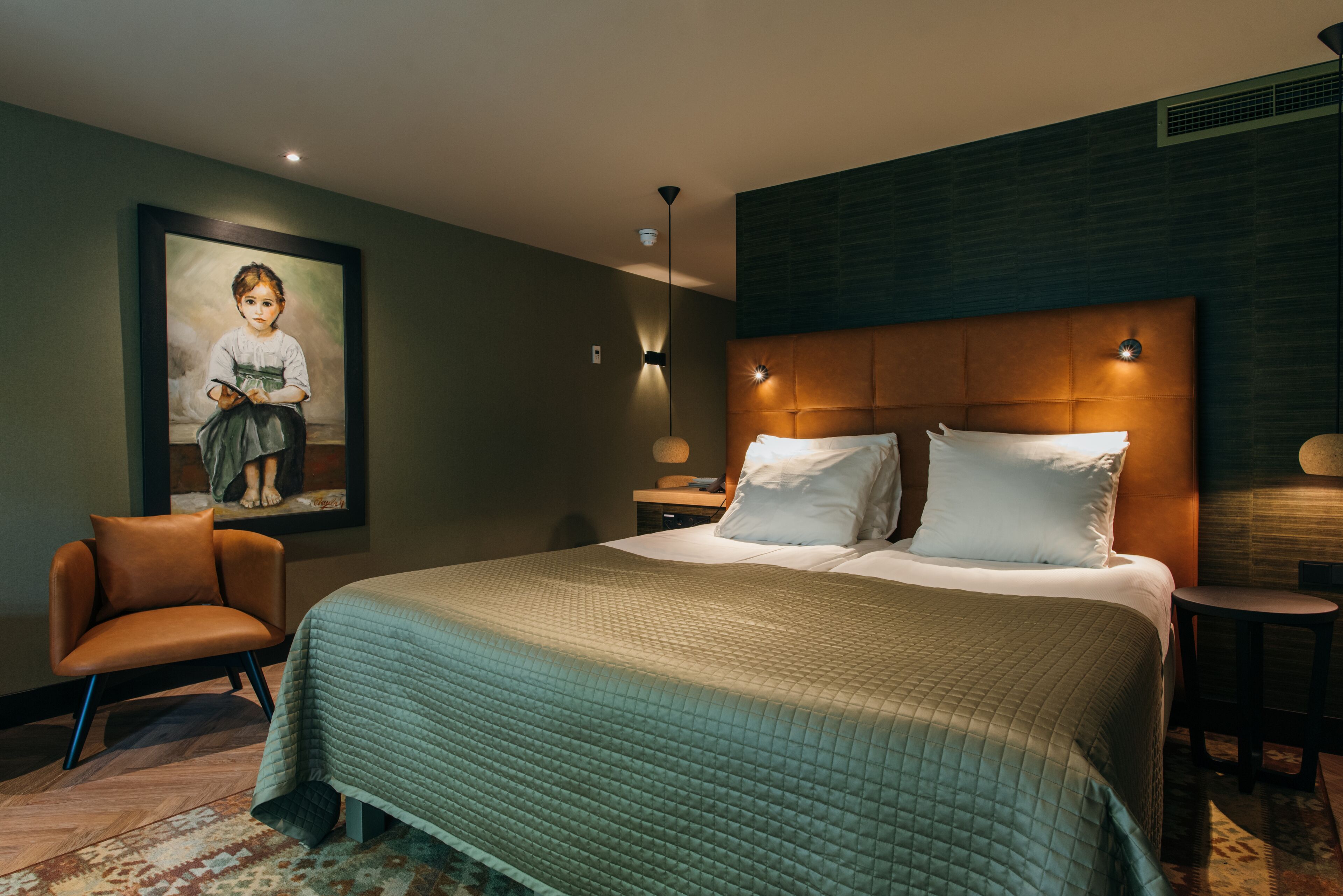 A hotel room with a large bed, a chair, a painting on the wall, and a nightstand with a lamp.