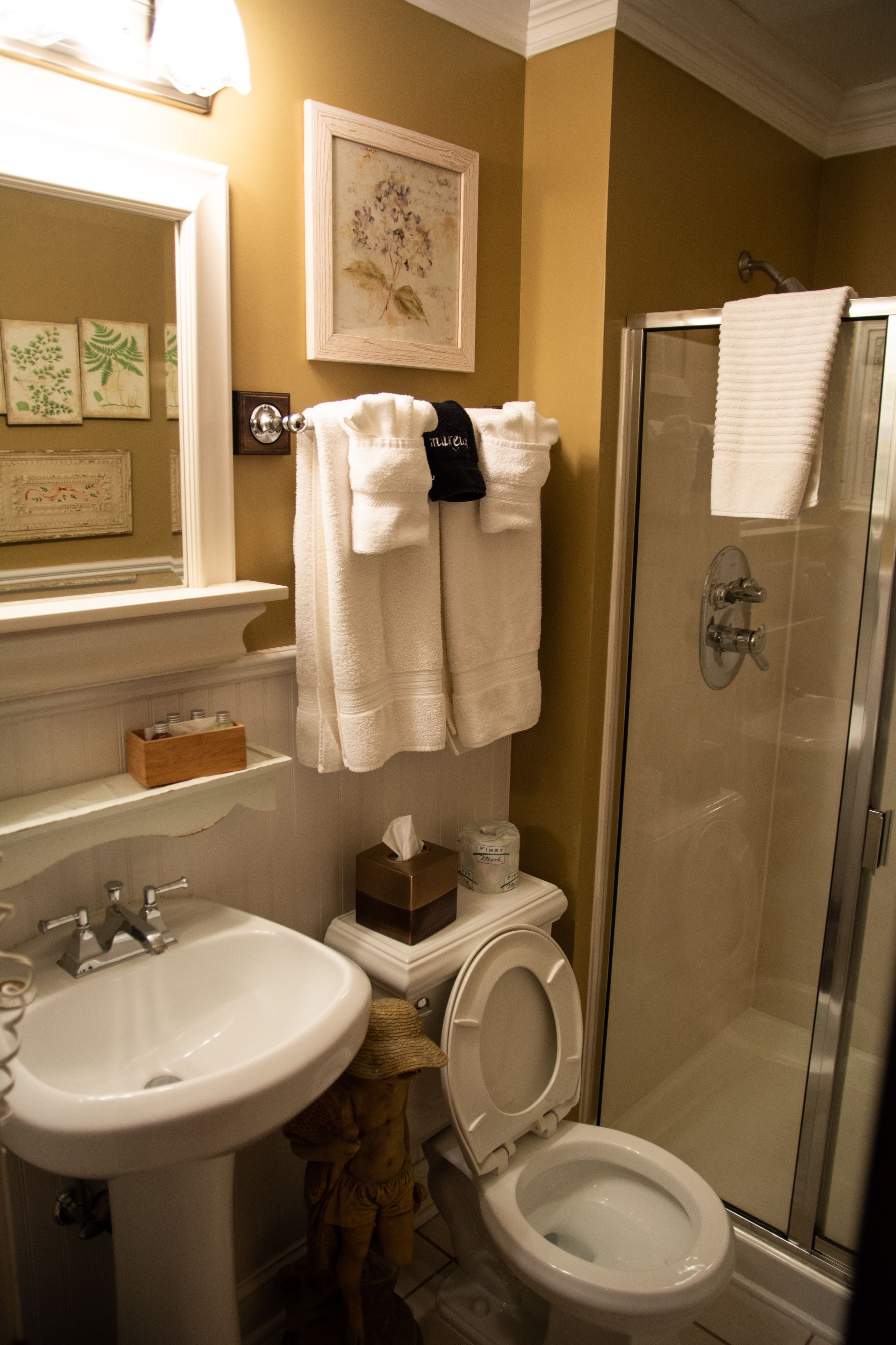 Un baño con lavamanos, inodoro y ducha con mampara de vidrio. Hay toallas colgadas en la pared y un cuadro enmarcado sobre el lavamanos.