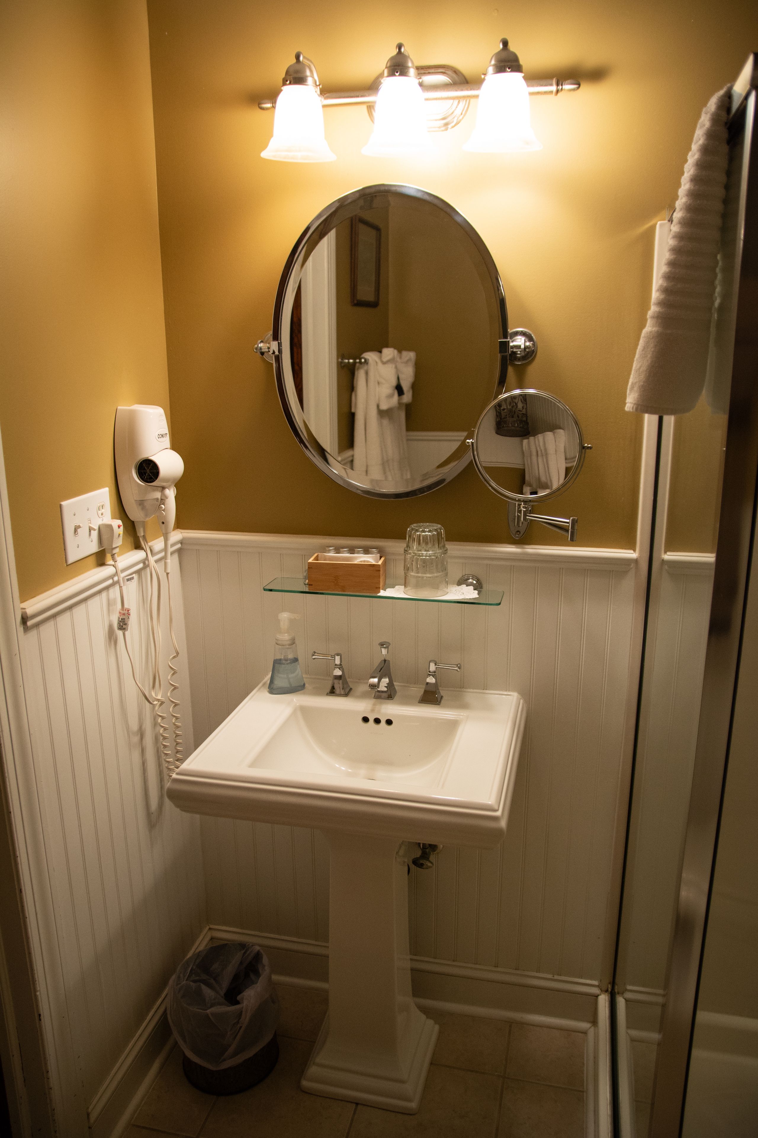 Baño con lavamanos blanco, dos espejos, secador de pelo y repisa de vidrio con artículos de aseo.