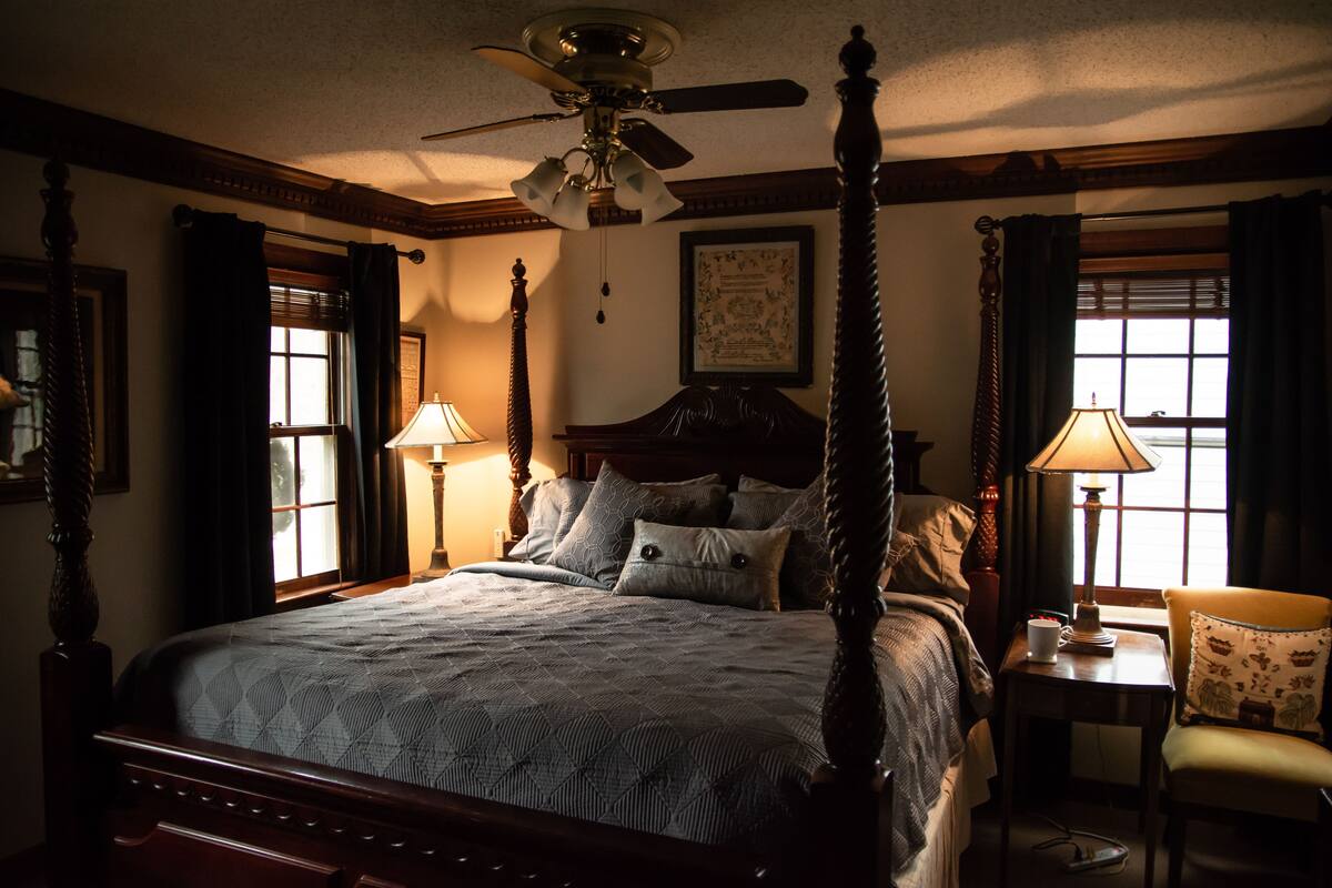 A four-poster bed with a quilt, two bedside lamps, a chair, and a ceiling fan in a room with windows and a framed picture on the wall.