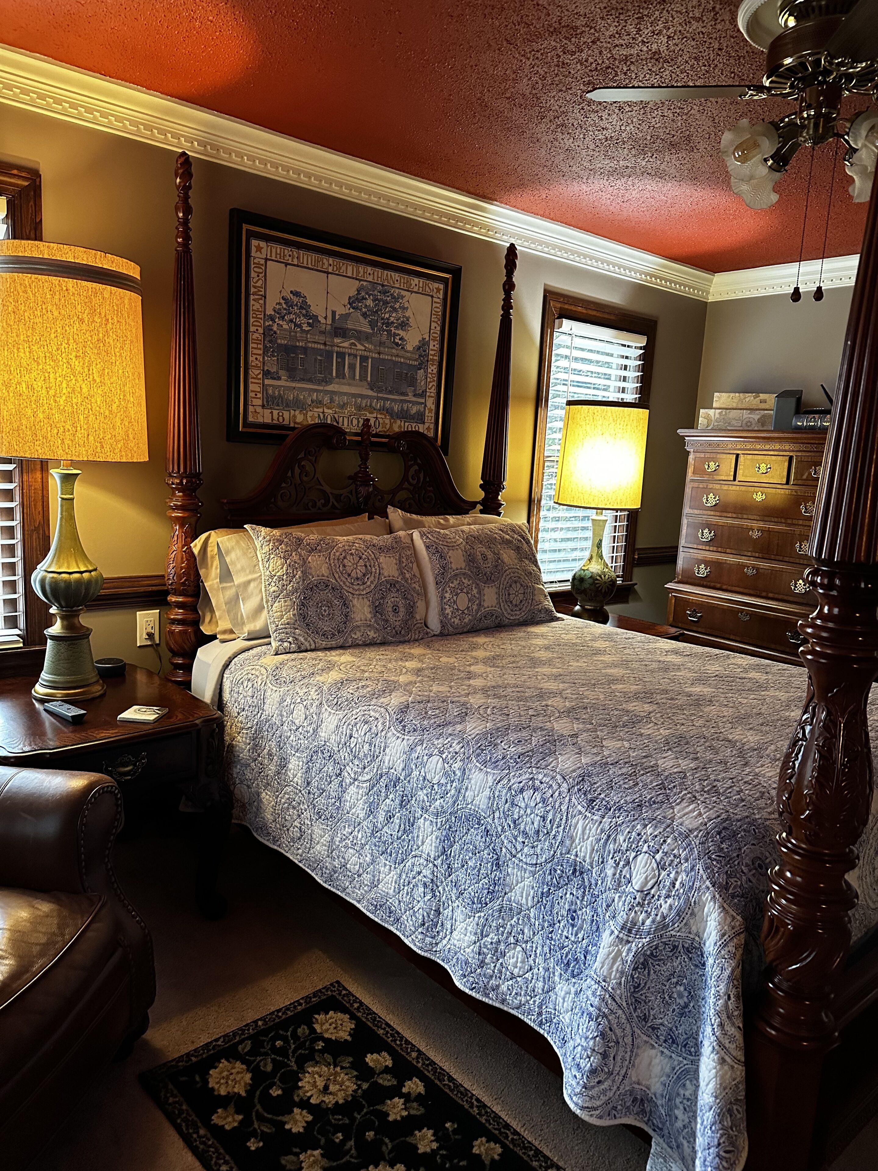 A bedroom with a four-poster bed, a nightstand with a lamp, a dresser, and a framed picture on the wall.