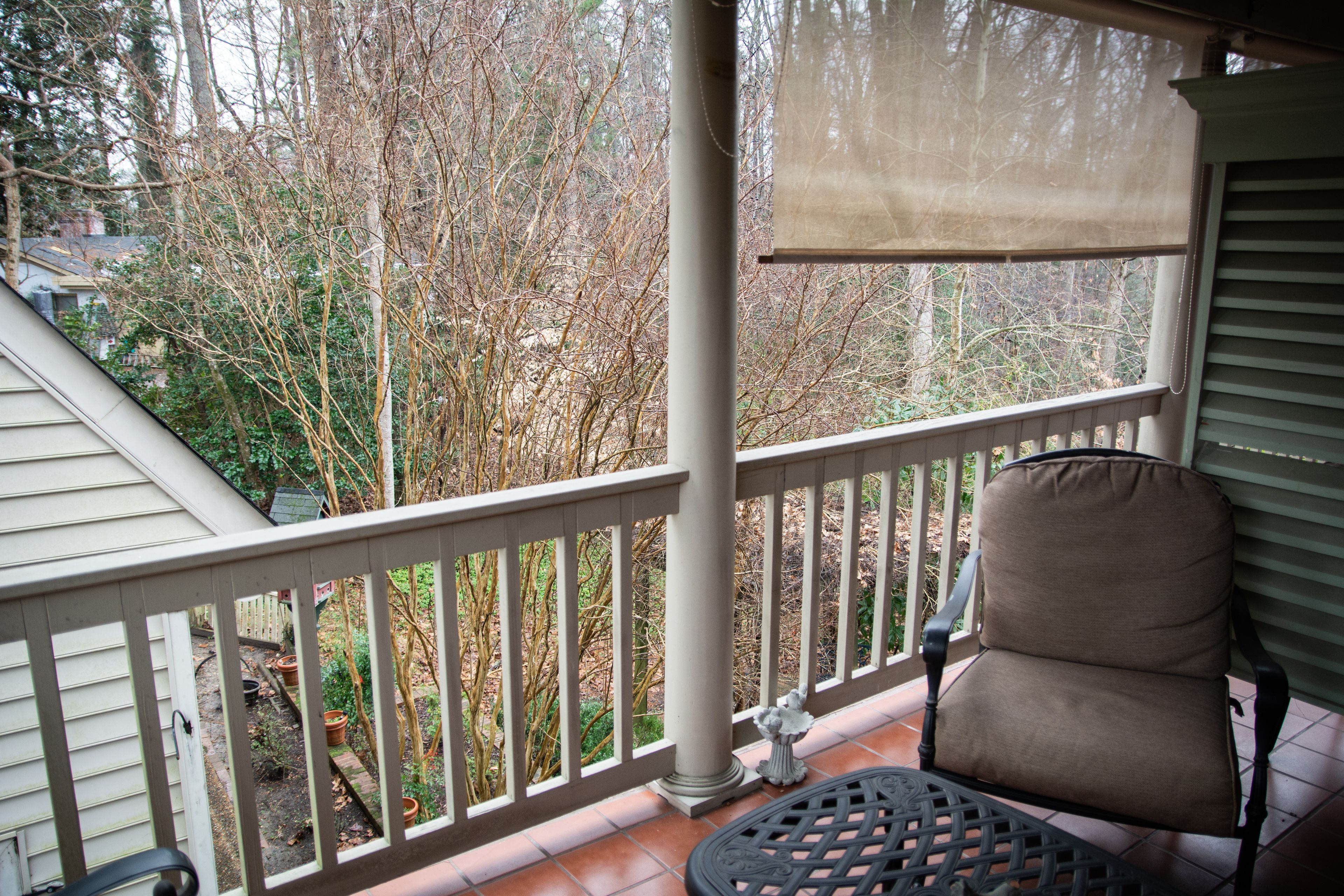 A porch with a chair and a table, a view of trees, and a house in the background.