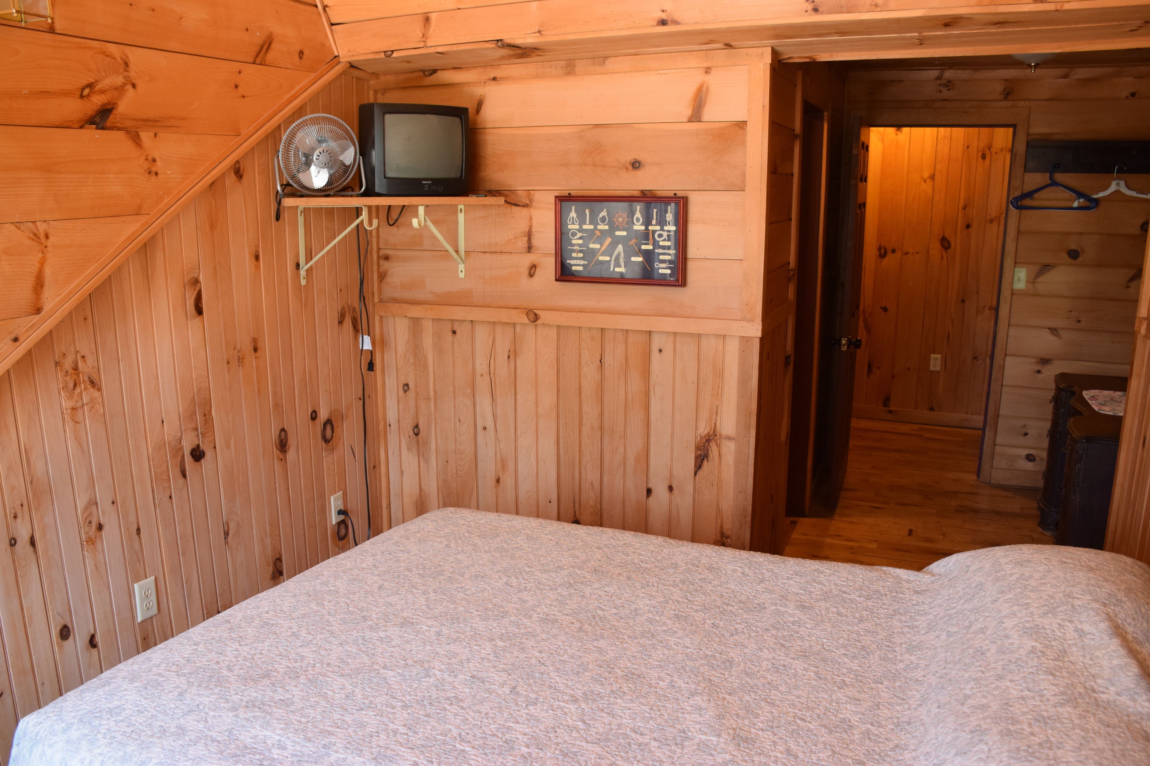 A wooden cabin bedroom with a bed, a wall-mounted TV, and a small shelf with a fan.