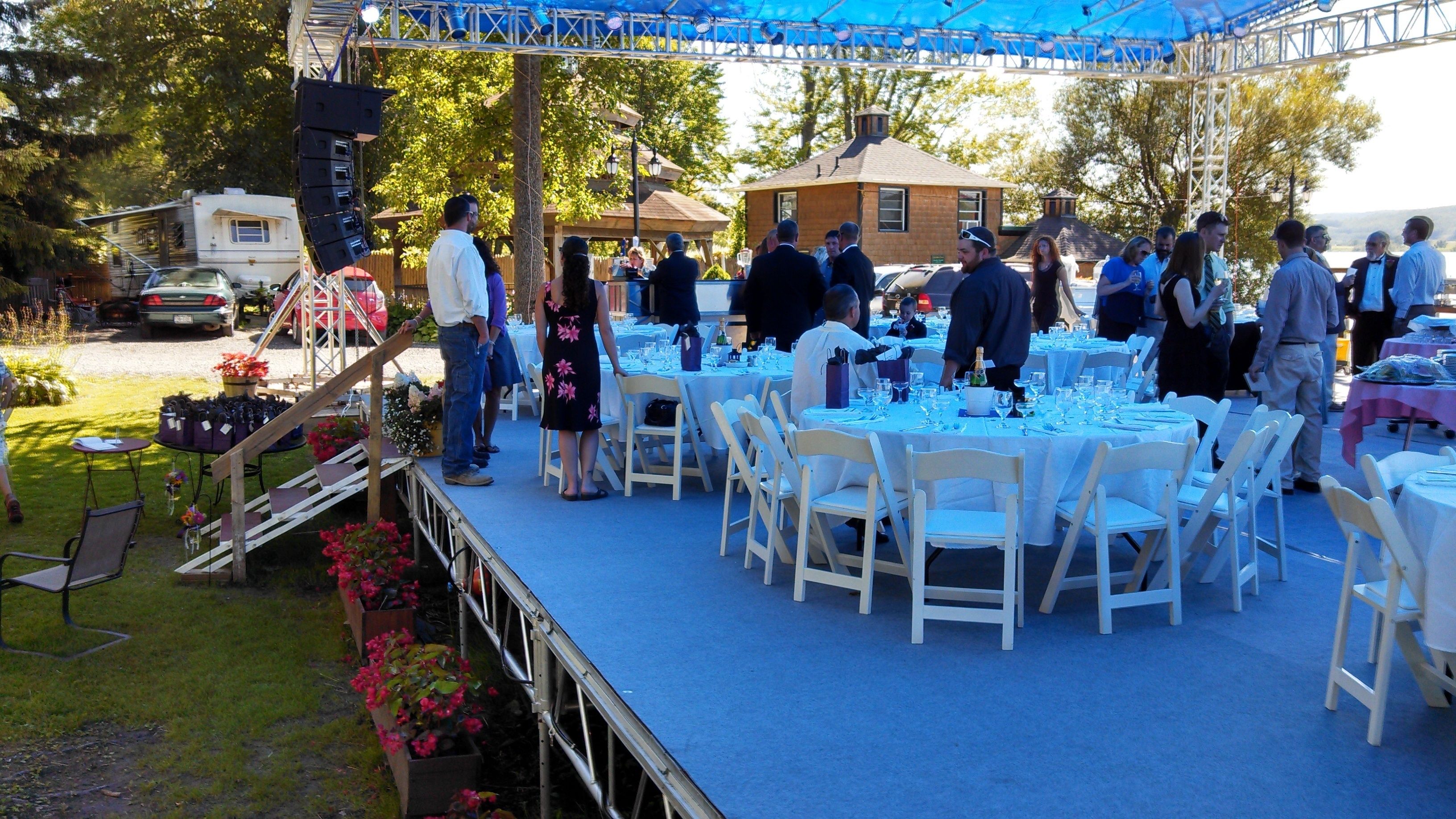 An outdoor event with round tables set up on a blue carpet, surrounded by white chairs.