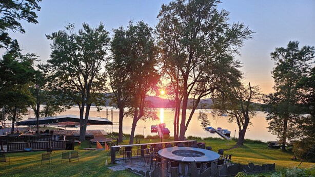 A lakeside outdoor seating area with a circular fire pit, picnic tables, and a covered pavilion.