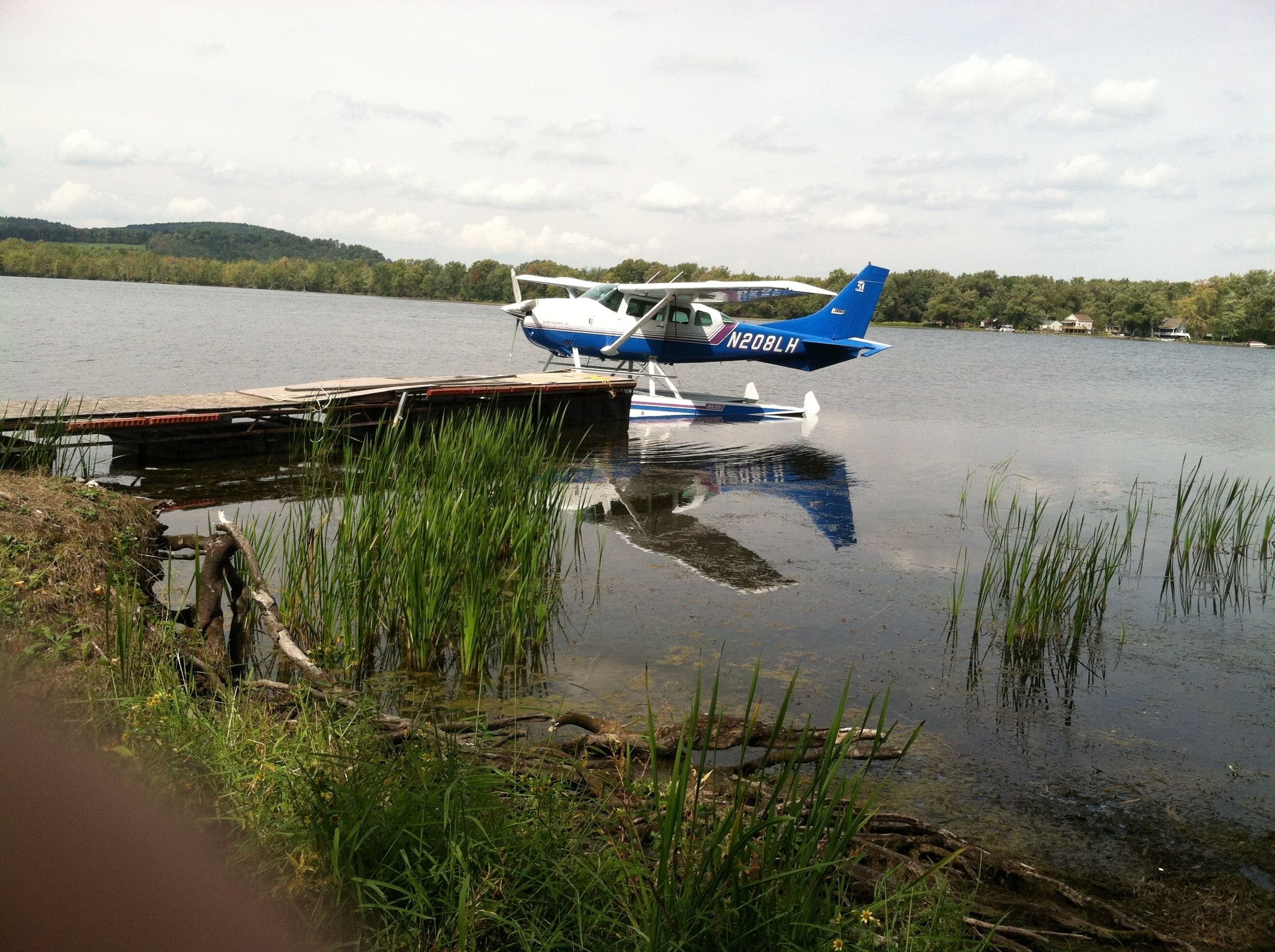 A small seaplane docked at a wooden pier on a calm lake with surrounding greenery.