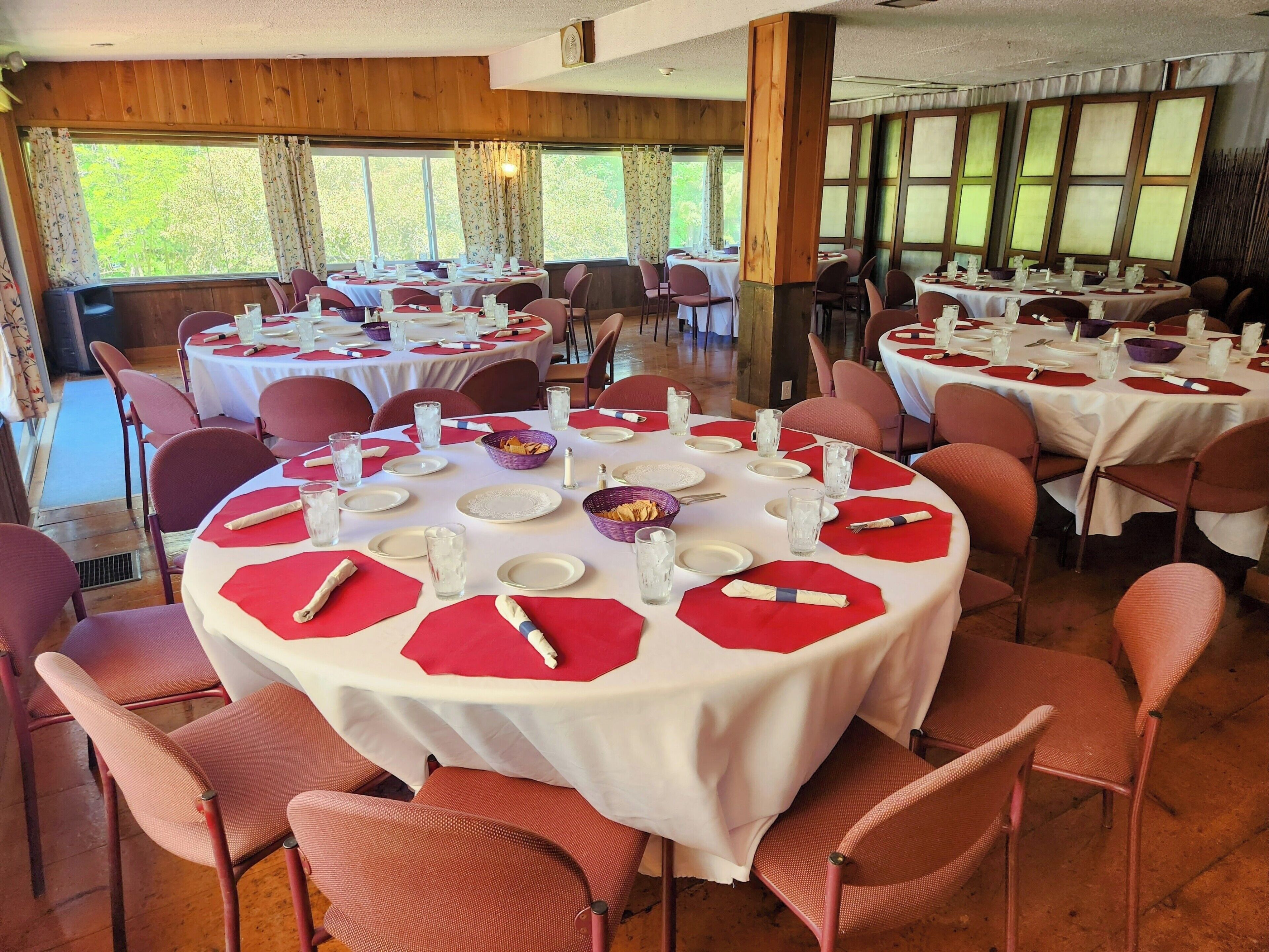 Comedor con mesas redondas dispuestas para varios comensales, con manteles blancos, posavasos rojos y cubiertos de metal.