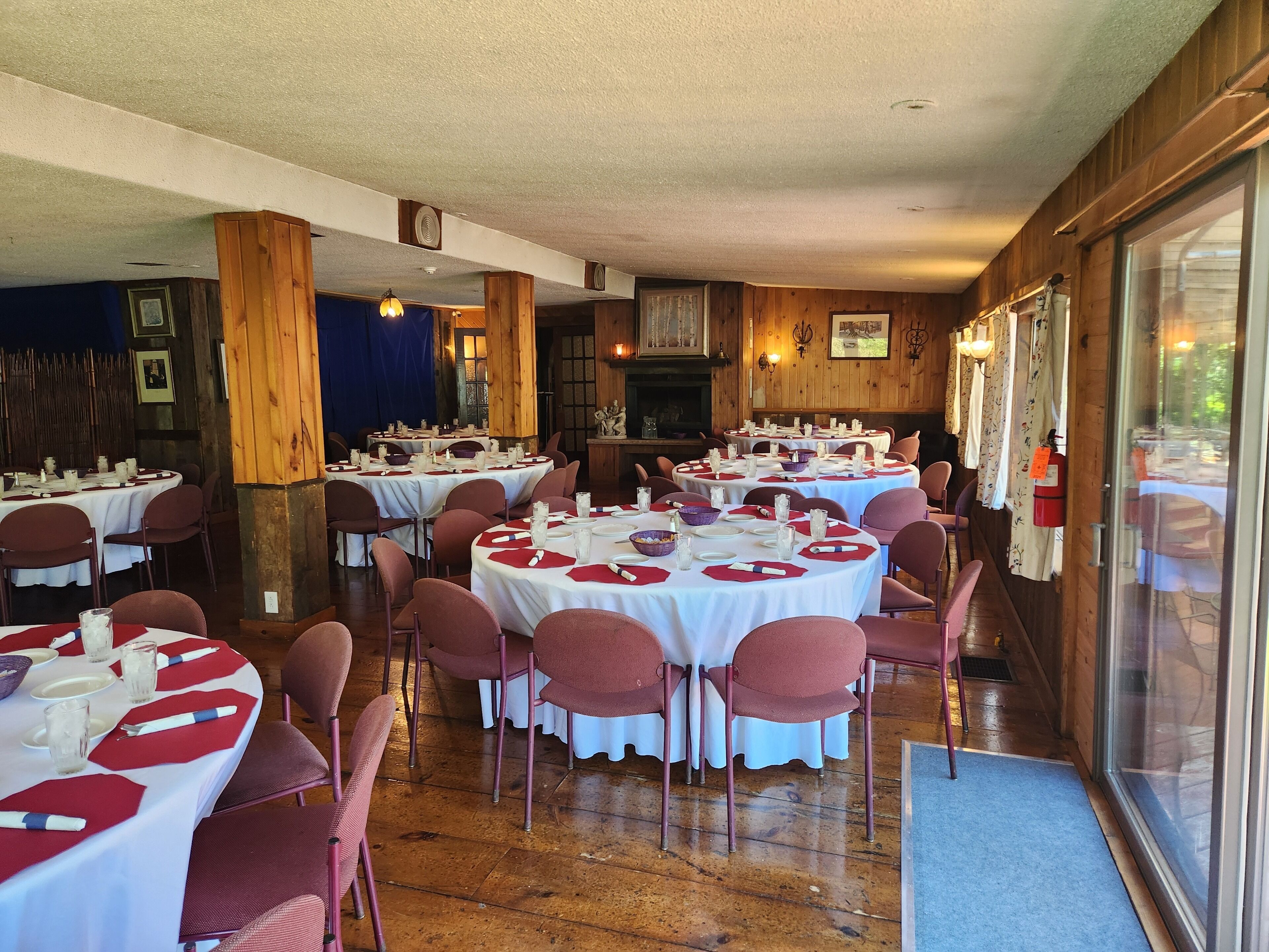 A restaurant with multiple round tables set with white tablecloths and red napkins, surrounded by wooden walls and large windows.