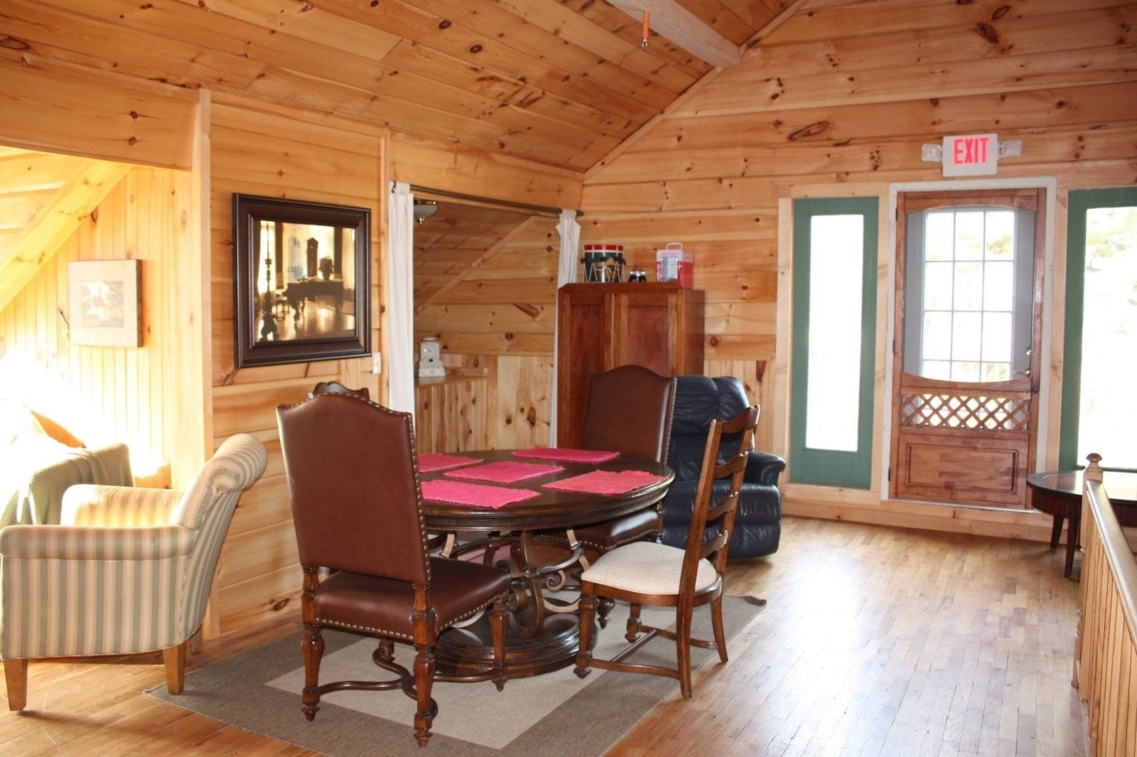 A wooden cabin interior with a dining table, chairs, a framed picture, and an exit sign.