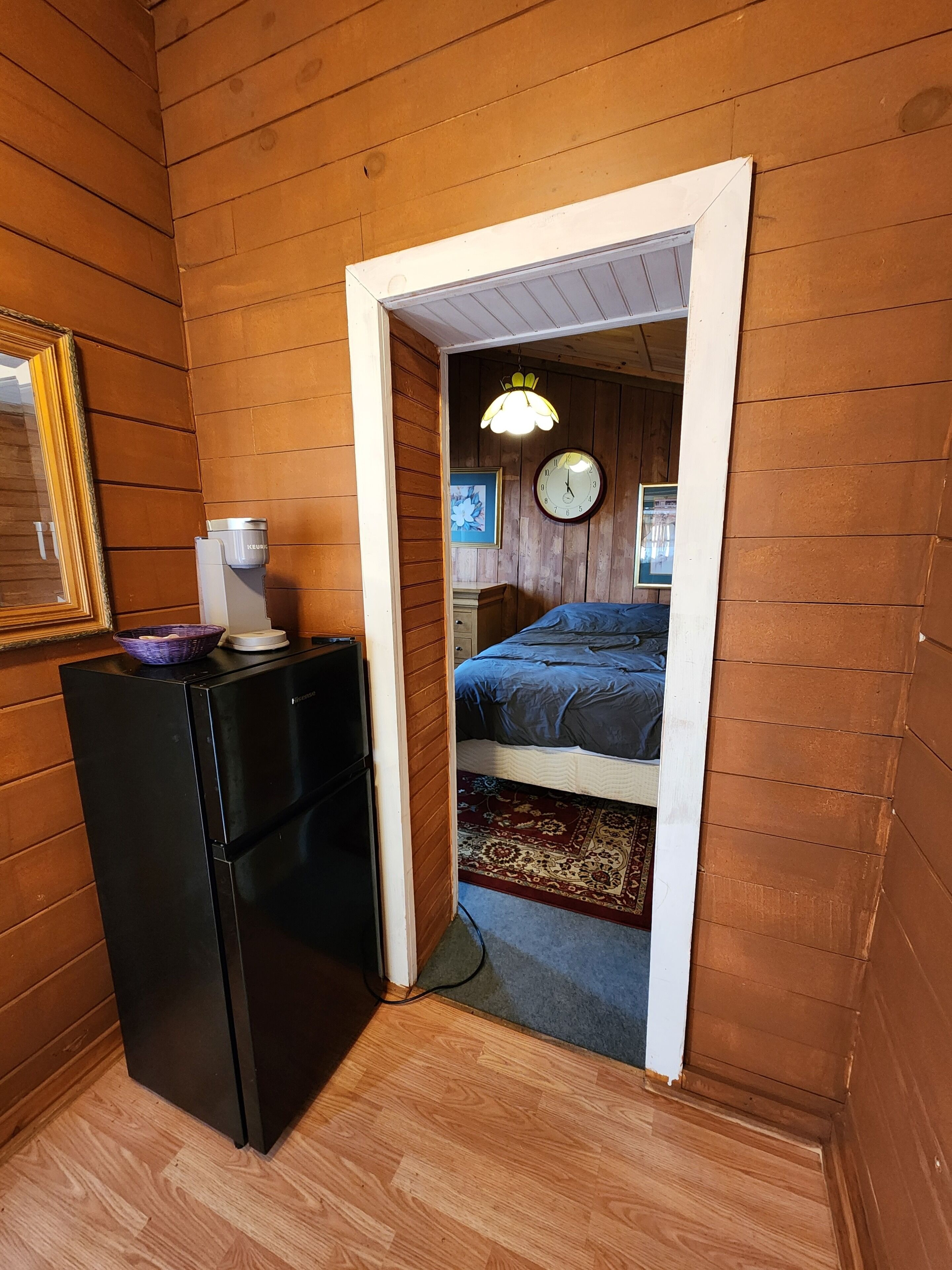 A compact room with wooden walls, a bed with a blue bedspread, a small refrigerator, and a wall clock.