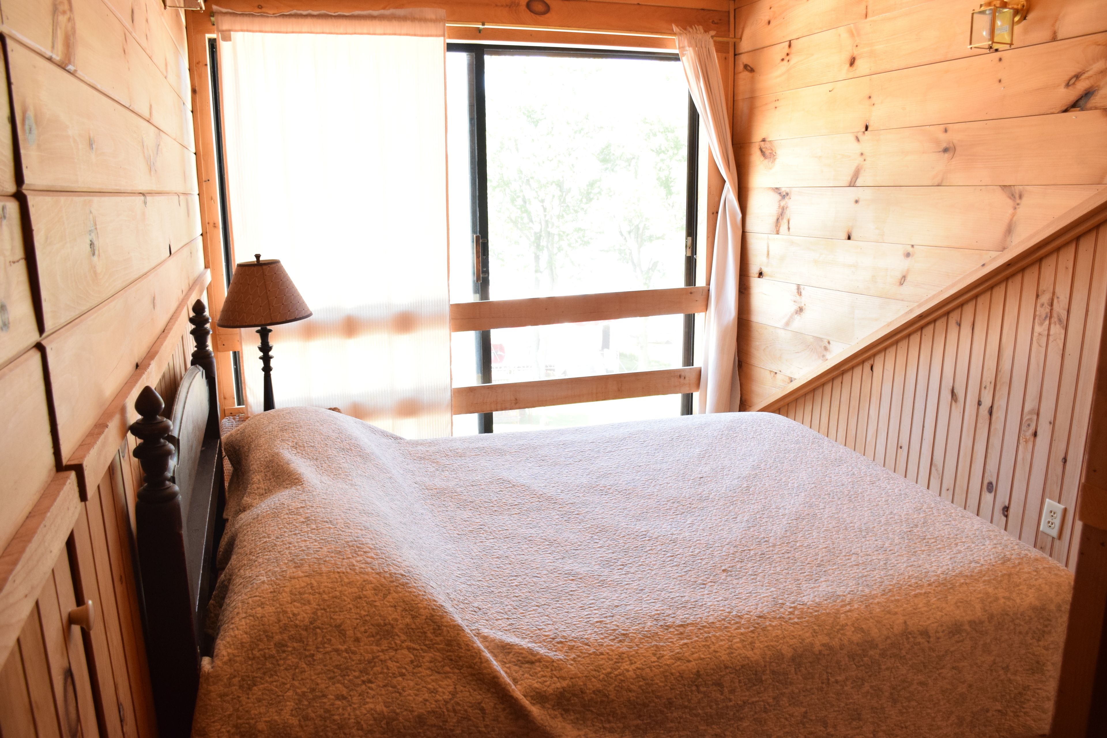 A wooden cabin room with a bed, a window, and a lamp.