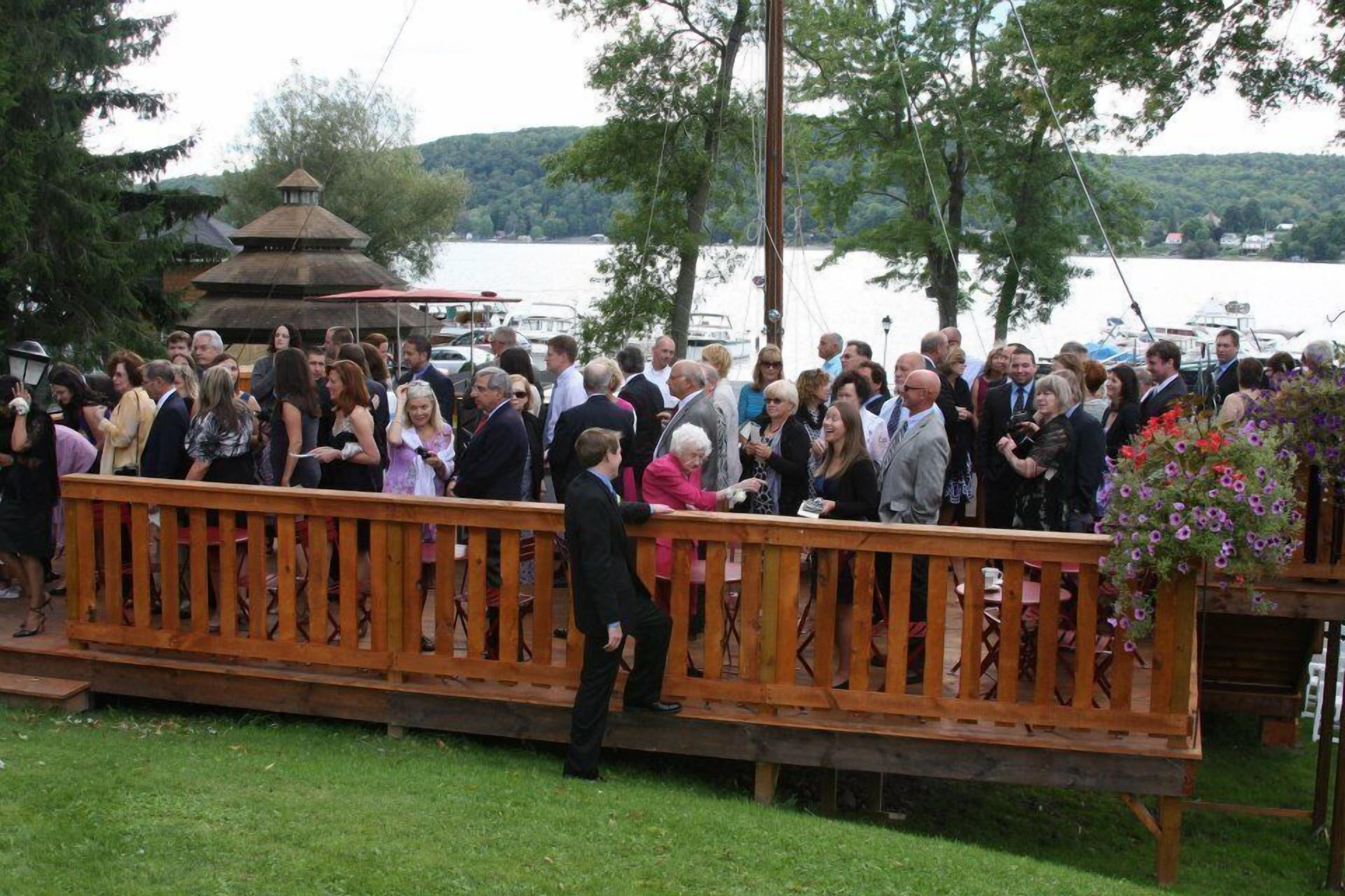 Un groupe de personnes s’est rassemblé sur une terrasse en bois, au bord d’un lac, avec des bateaux en arrière-plan.