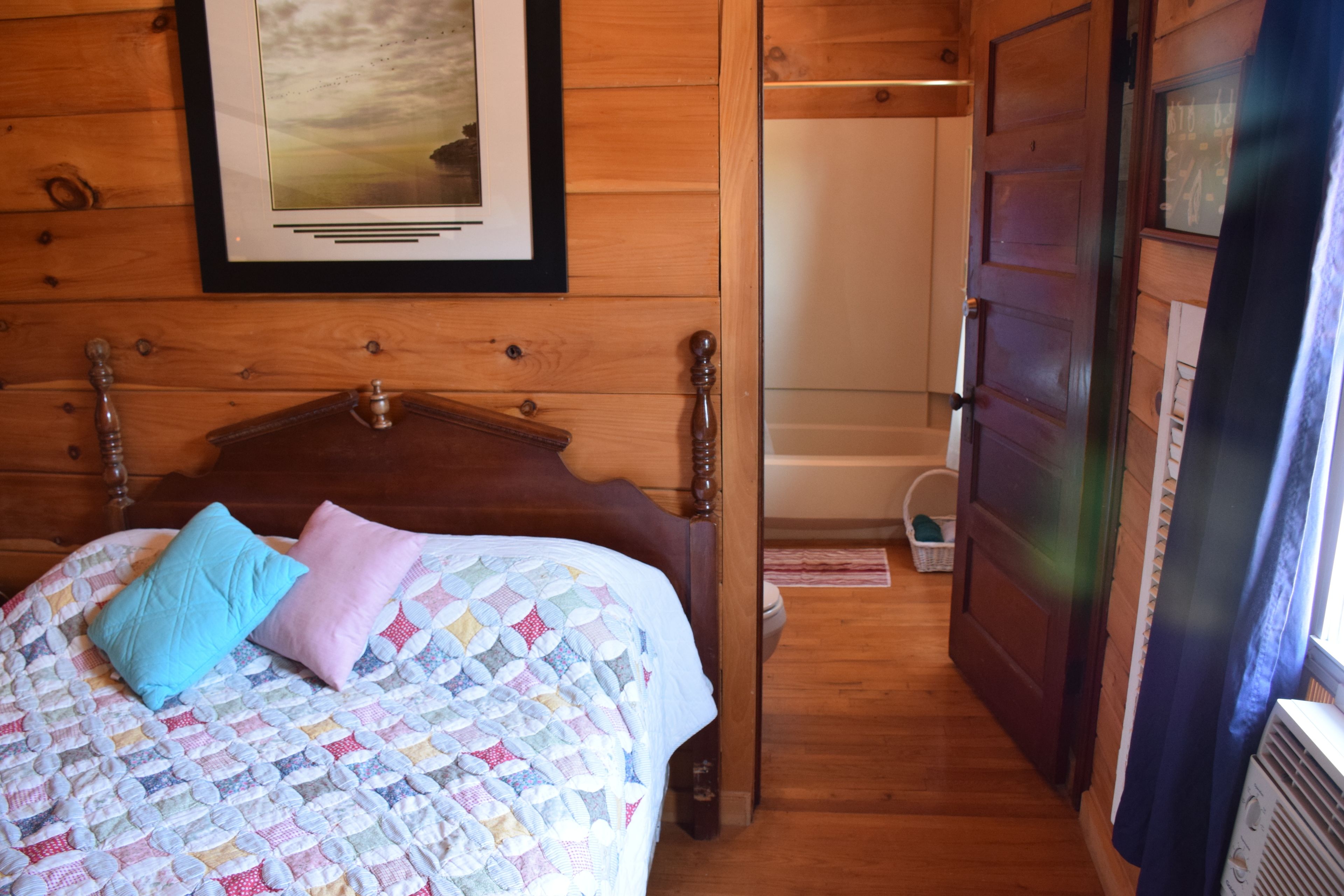 Una habitación con paneles de madera, una cama, un cuadro en la pared y una puerta que da a otra habitación.