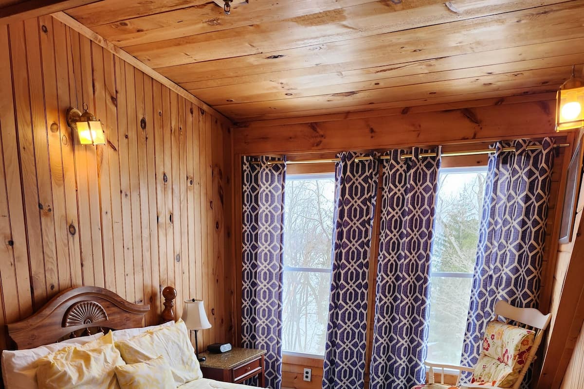 Ein Schlafzimmer einer Holzhütte mit einem großen Bett, gemusterten Vorhängen und Blick auf Bäume.
