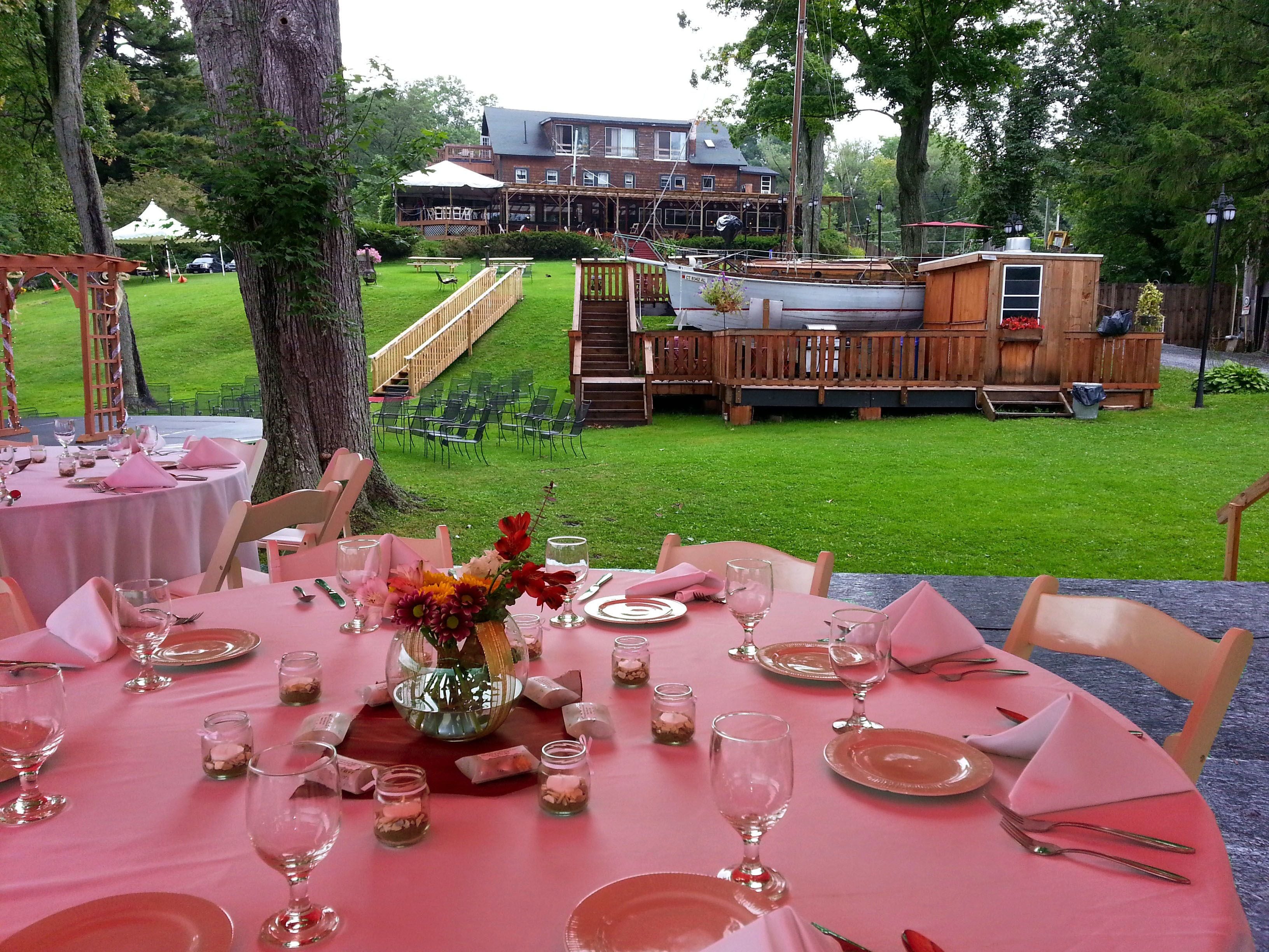 A dining area with pink tablecloths and glassware, set against a backdrop of a wooden deck and a building with a covered patio.