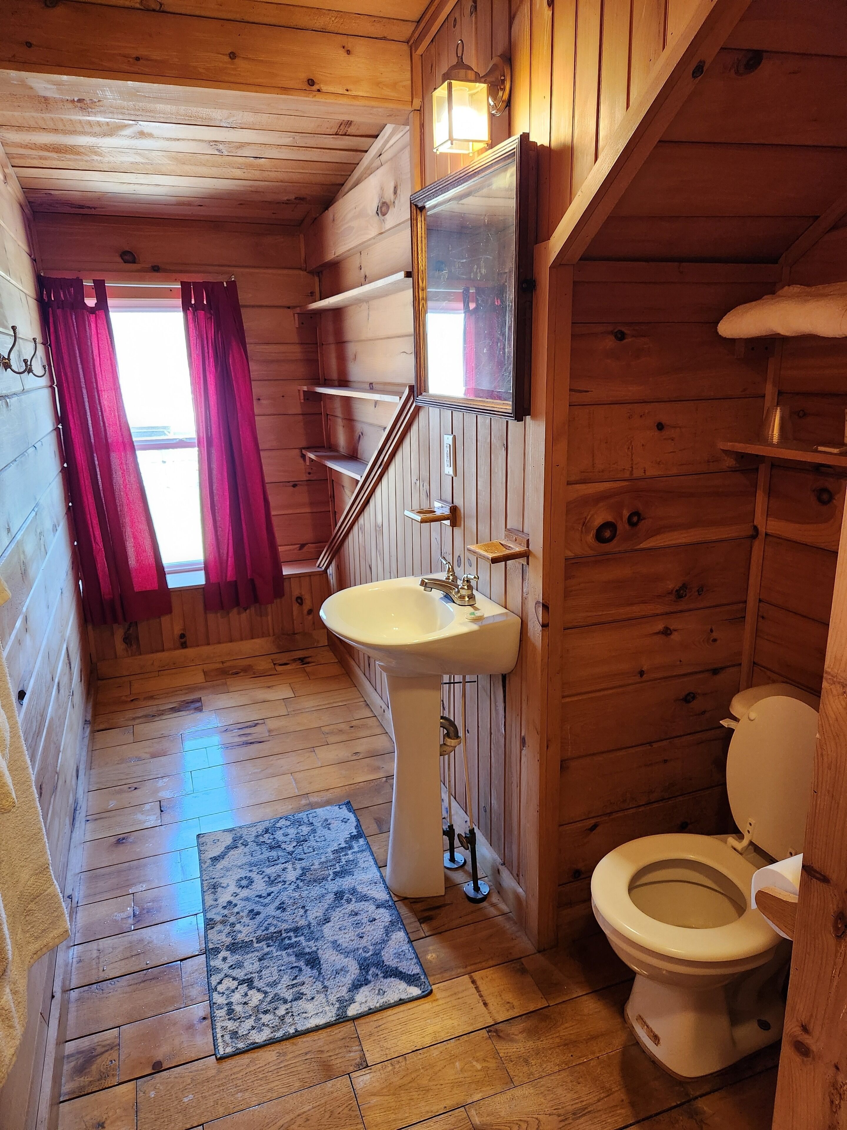 Bagno di una baita in legno con lavandino, WC e finestra con tende rosse.