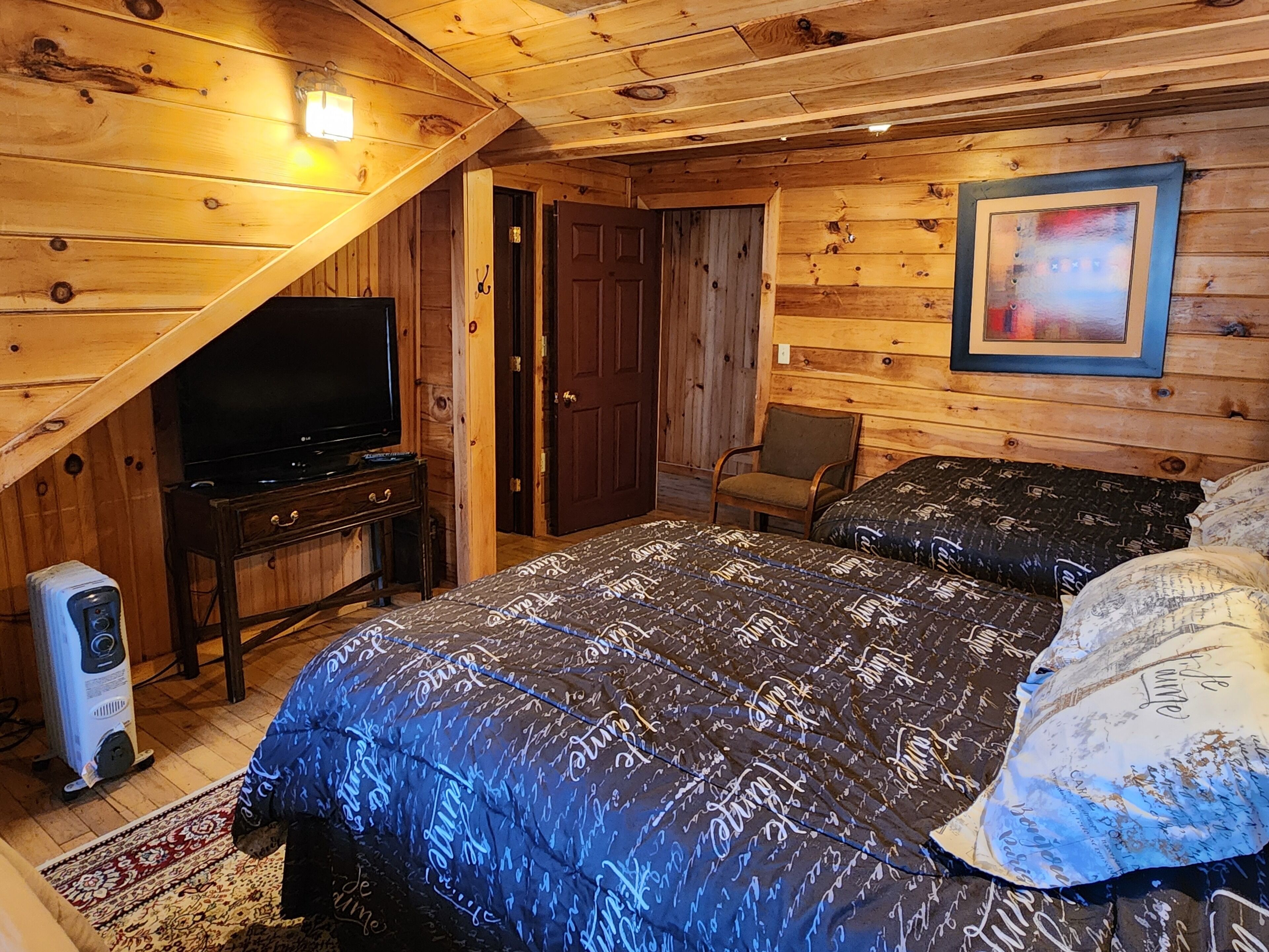 A cozy log cabin bedroom with a large bed, a television on a wooden stand, a chair, and a framed picture on the wall.
