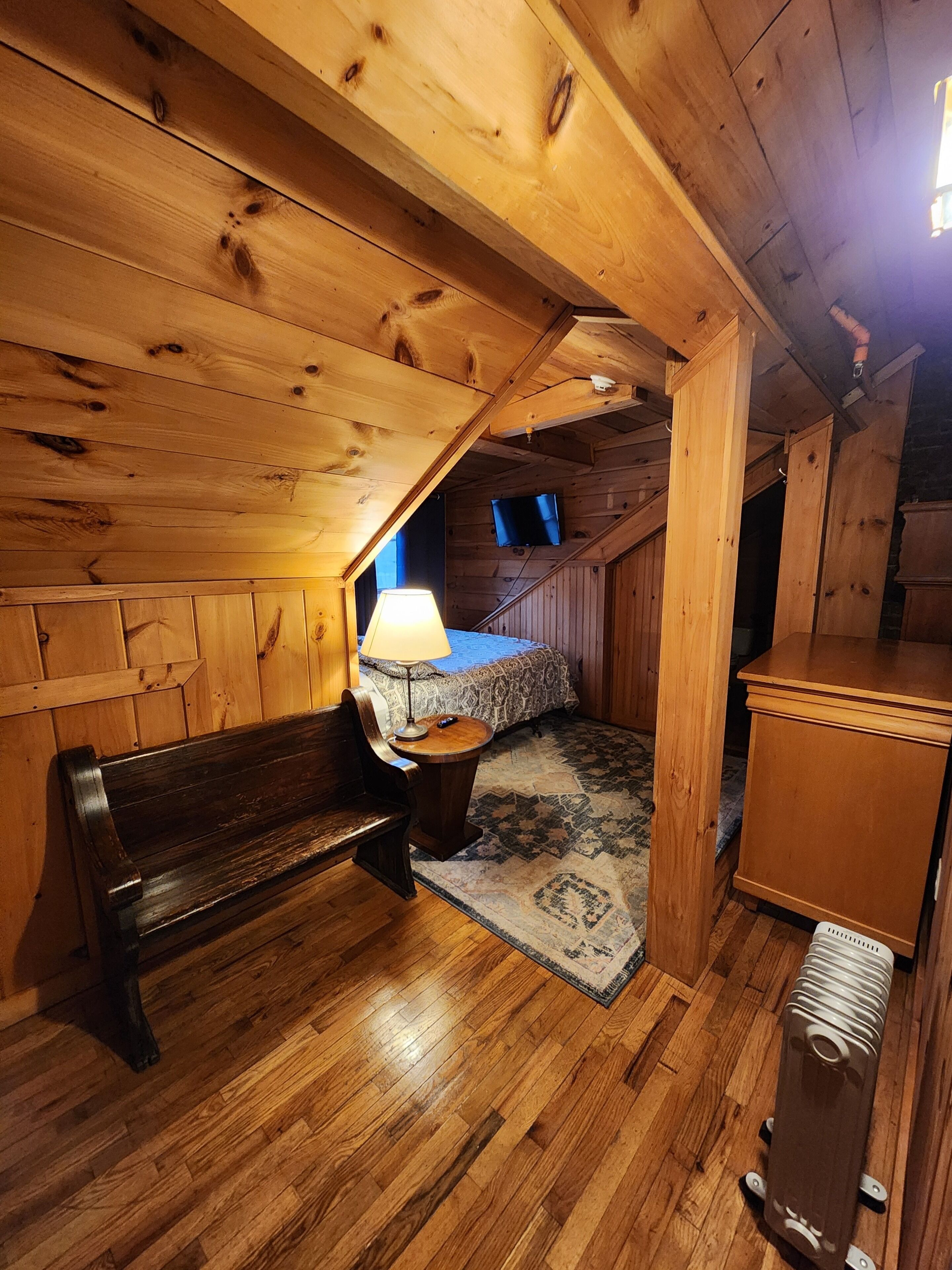 A cozy attic bedroom with wooden walls and a sloped ceiling, featuring a bed, a bench, a small table with a lamp, and a radiator.