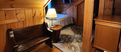 A cozy attic bedroom with wooden walls and a sloped ceiling, featuring a bed, a bench, a small table with a lamp, and a radiator.