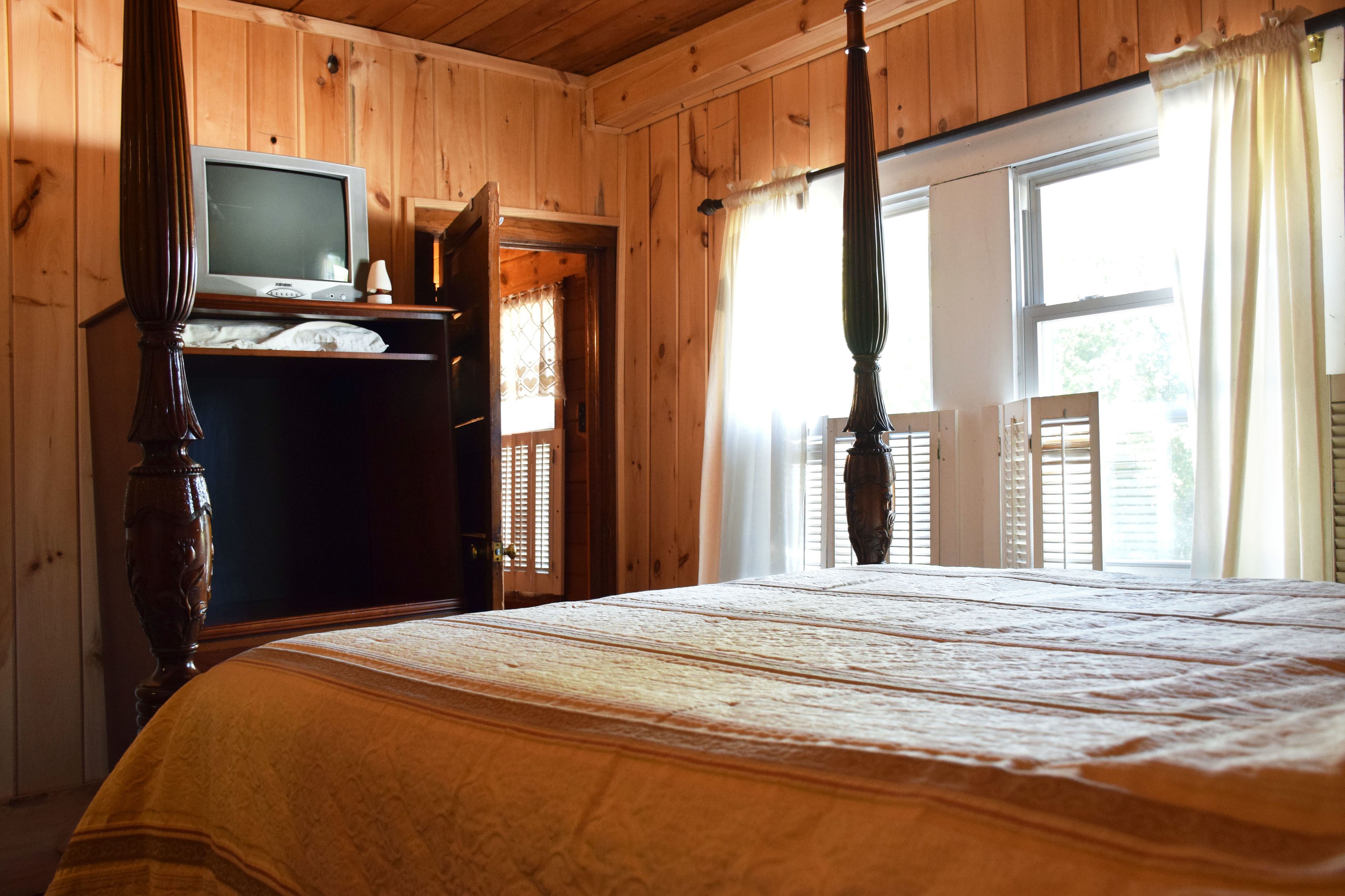 A wooden-paneled bedroom with a bed, a television on a dresser, and a window with shutters.