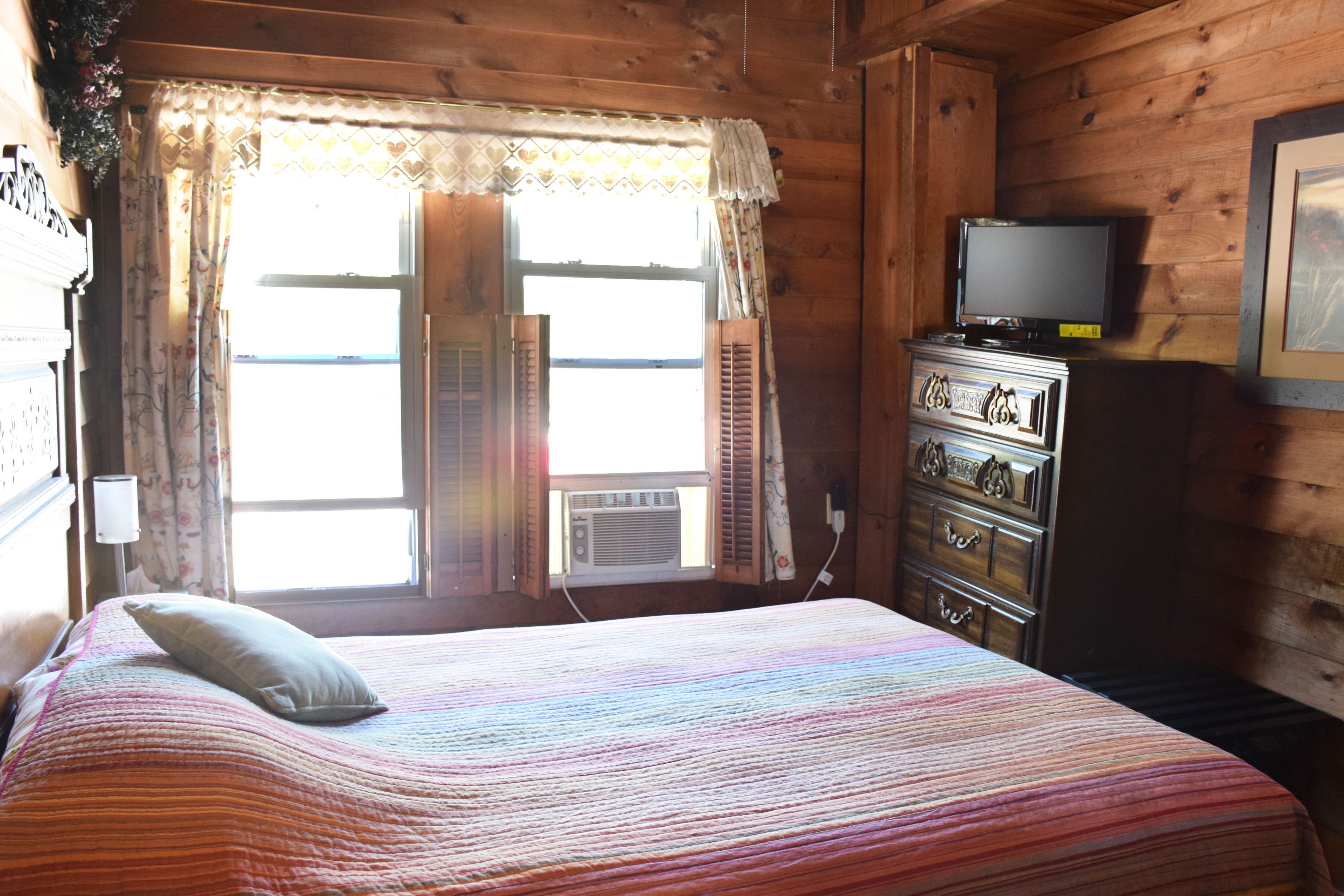 Una habitación de cabaña de madera con una cama, un aparador, un televisor y una ventana con contraventanas.
