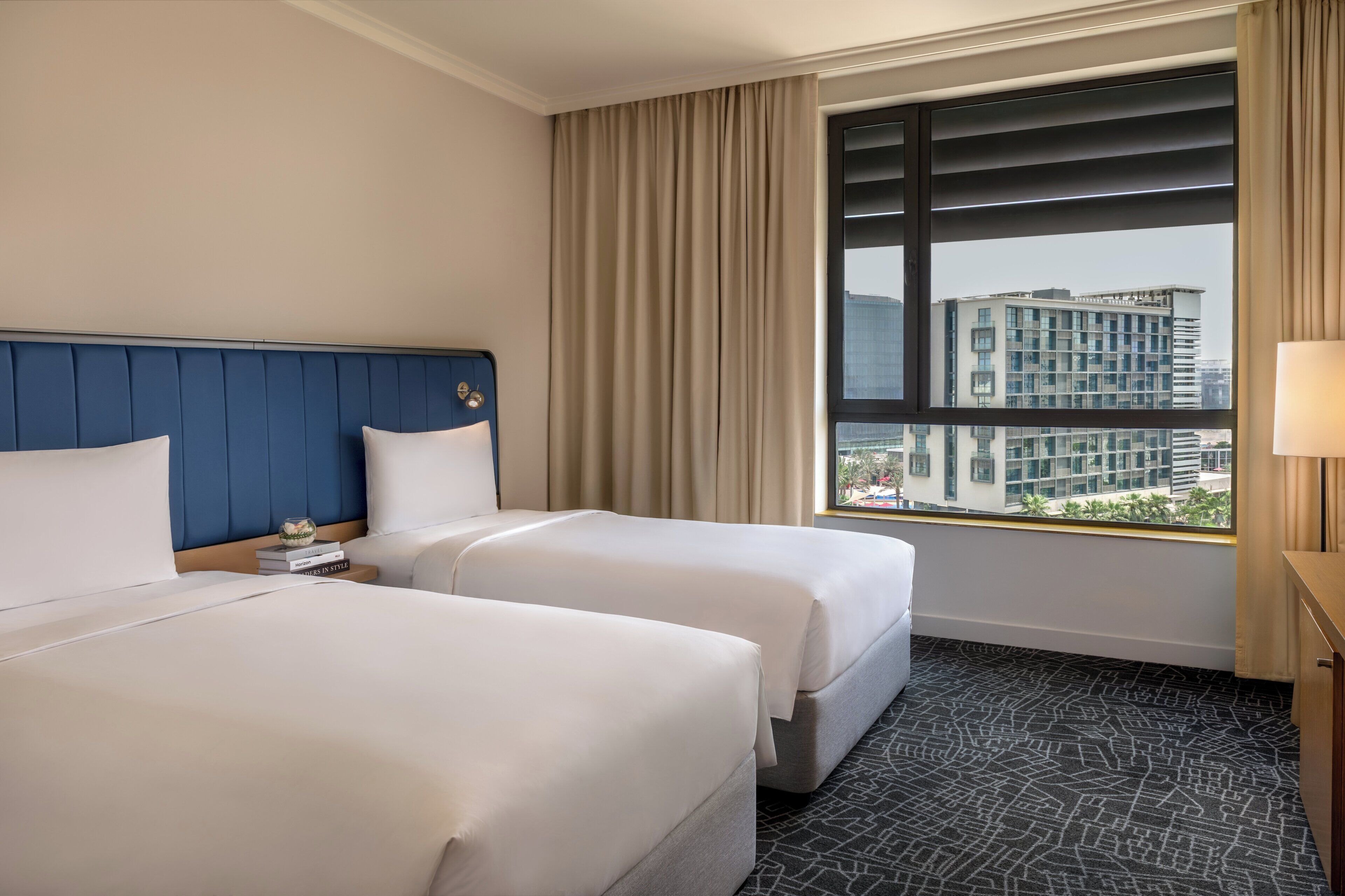 Quarto de hotel com duas camas, uma janela grande com vista para prédios e uma mesa de cabeceira com abajur.