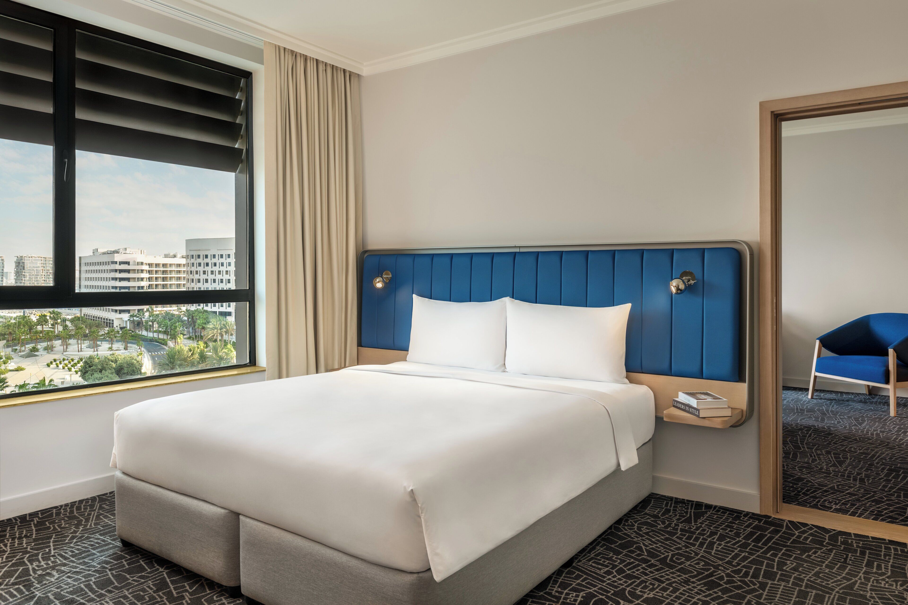 A hotel room with a large bed, a view of the city, and a blue chair.
