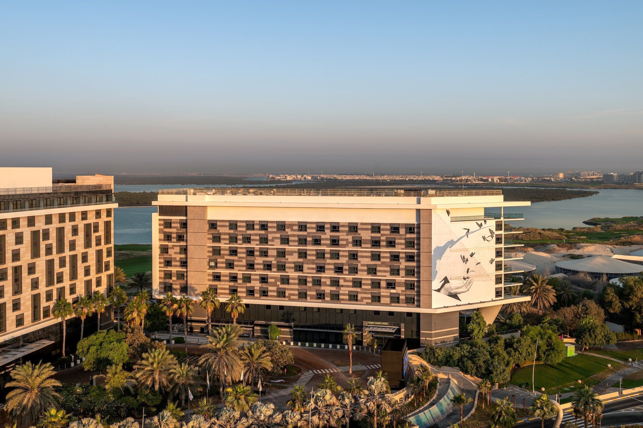 A modern hotel with a central courtyard, surrounded by palm trees and a view of a body of water.