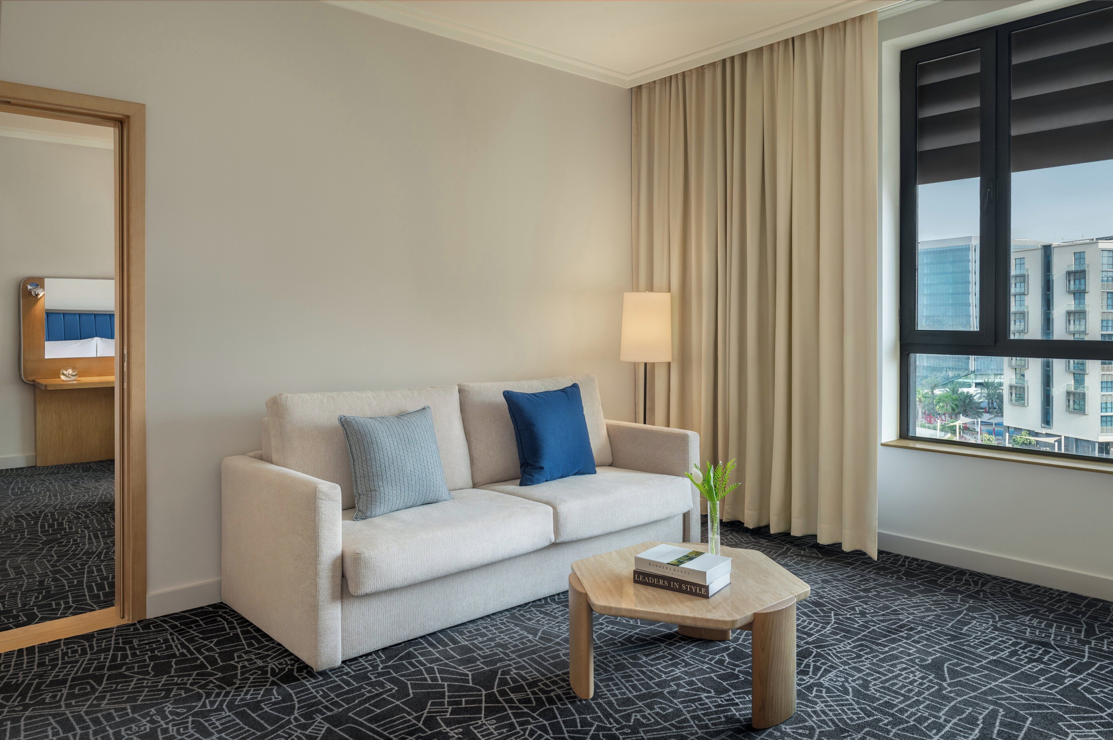 A hotel room with a sofa, a small table with a book, a lamp, and a window with a view of buildings.