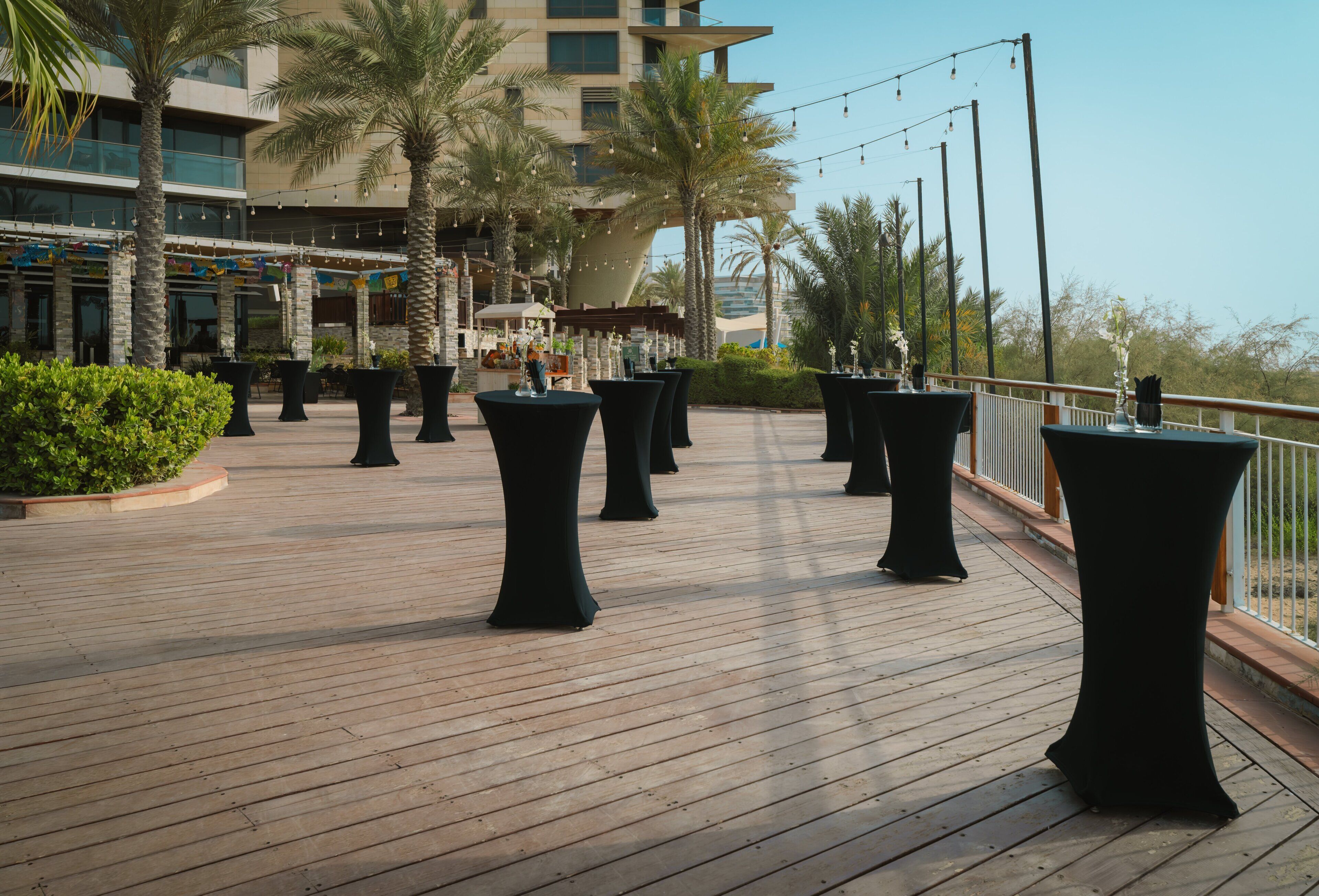 Una terraza de madera con mesas y sillas redondas, palmeras y un edificio con balcones al fondo.