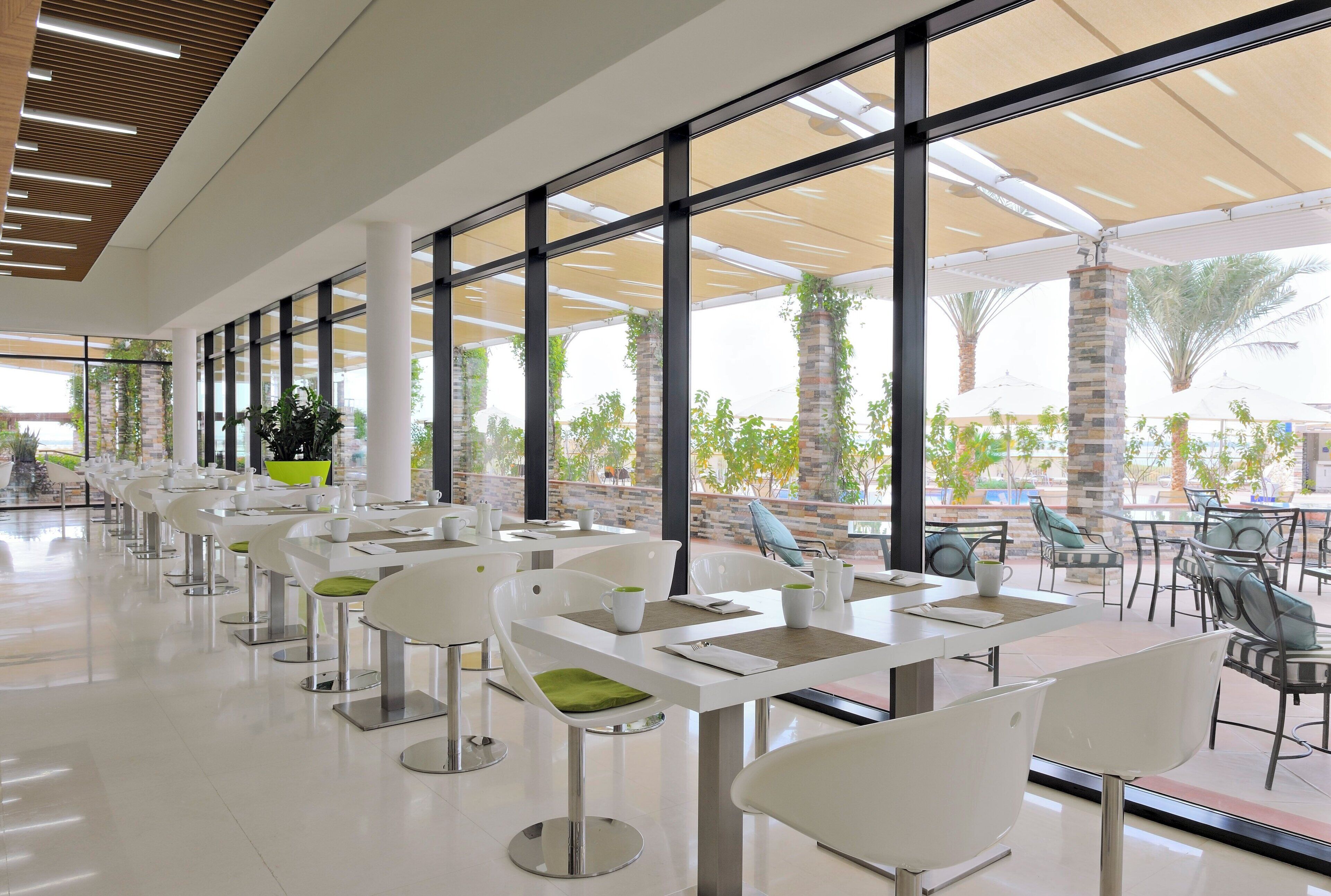 A modern indoor dining area with white tables and chairs, large windows, and a view of the outdoors.