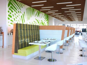 Un restaurante moderno con una barra larga, sillas blancas y una pared con estampado verde y blanco.