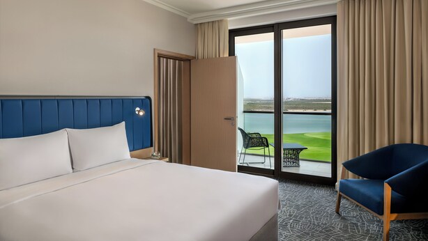 Habitación de hotel con una cama grande, cabecera azul, puerta de madera y vista a un campo de golf a través de una puerta corrediza de cristal.