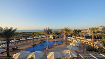 Ein Poolbereich eines Resorts mit Sonnenschirmen und Liegestühlen, umgeben von Palmen und Blick auf das Meer.