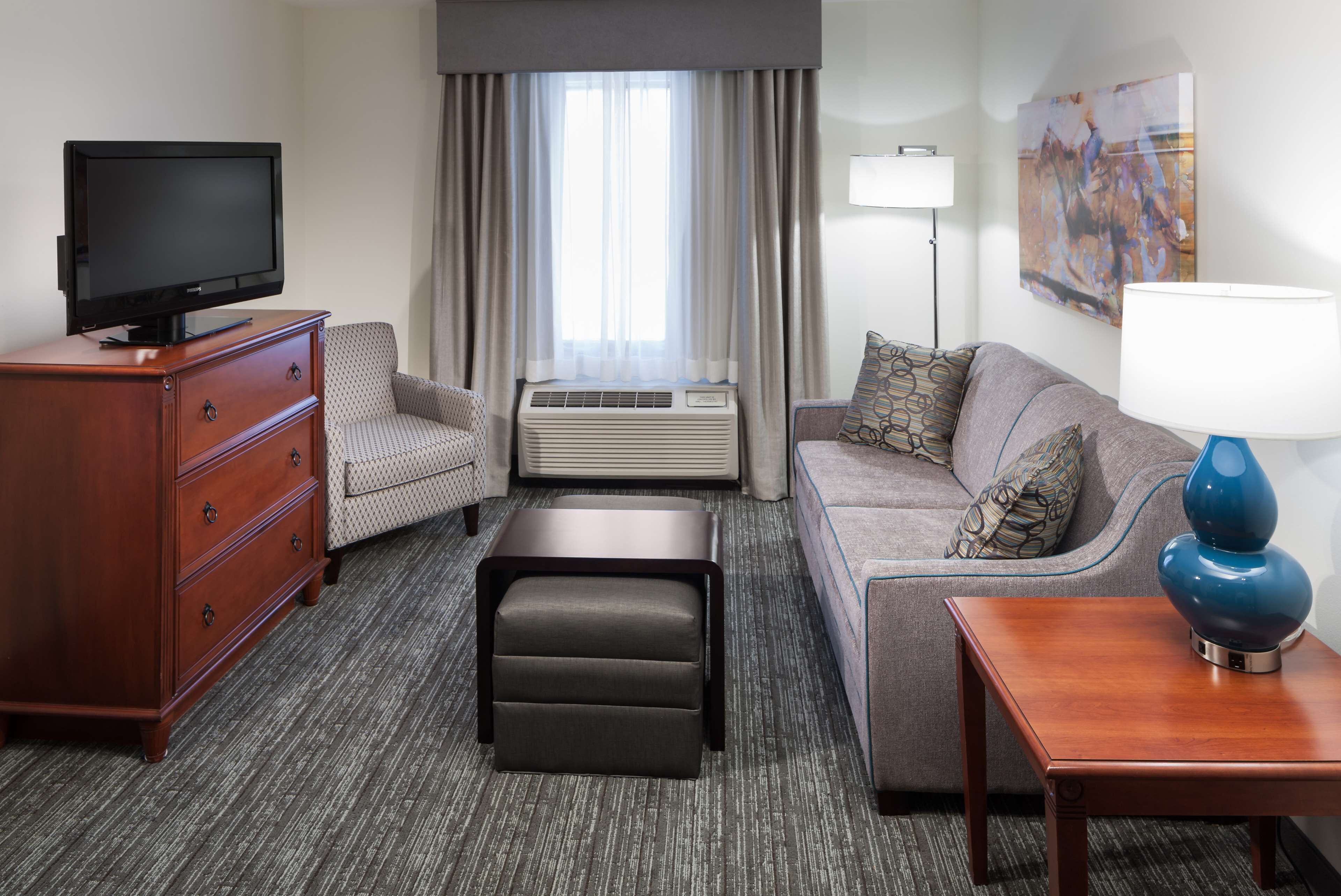A hotel room with a flat-screen TV on a wooden dresser, a sofa with patterned cushions, a small table with a lamp, and a large window with curtains.