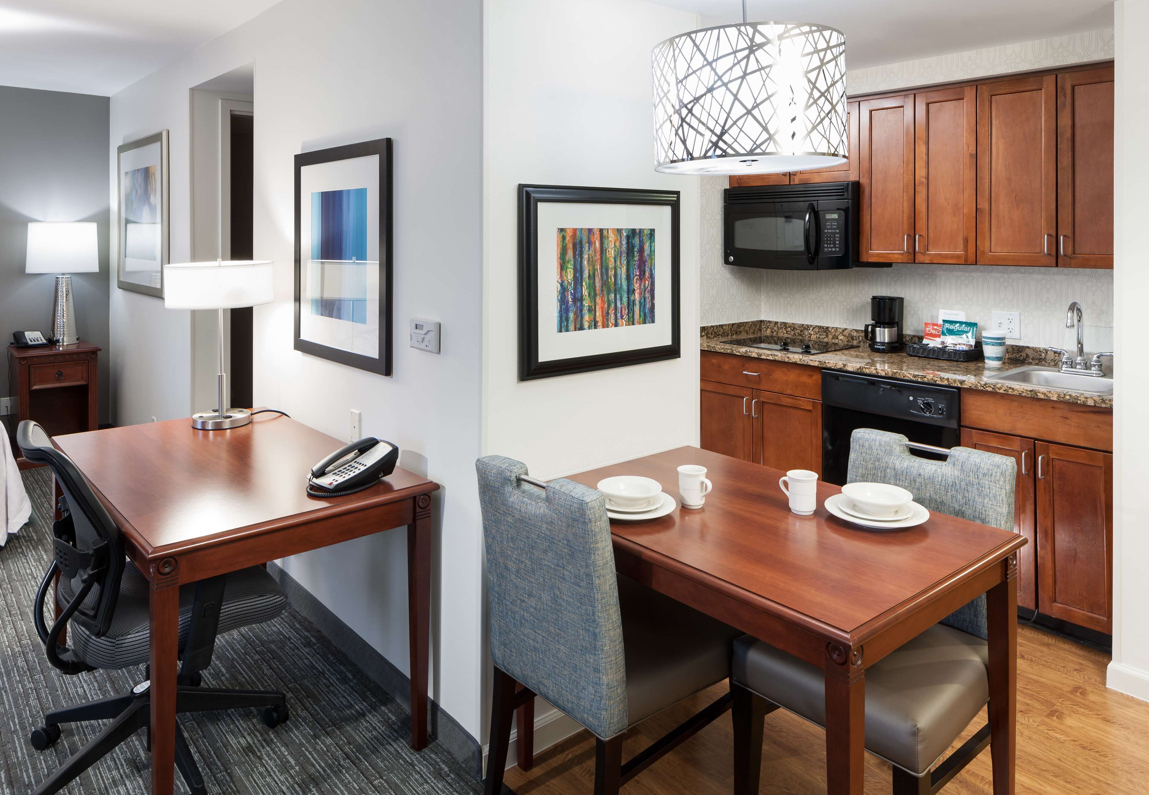 A hotel room with a wooden dining table, two chairs, a desk with a telephone, a lamp, framed artwork, a microwave, a coffee maker, and a sink.