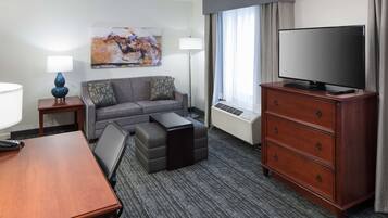 A hotel room with a grey sofa, a wooden dresser, a flat-screen TV, a lamp, and a painting on the wall.