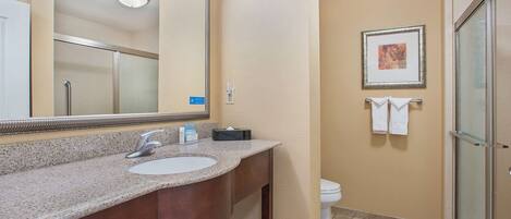 A bathroom with a granite countertop sink, a large mirror, a framed picture on the wall, and a glass shower enclosure.
