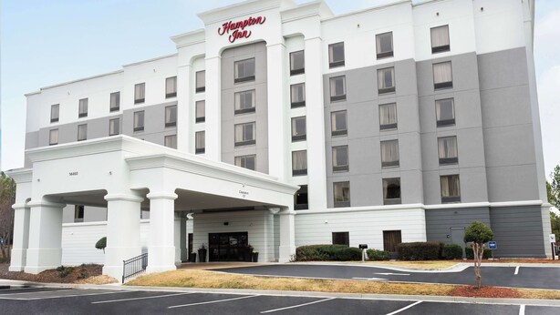 A large, multi-story hotel with a prominent 'Hampton Inn' sign. It features a central entrance with a covered area and multiple windows on each floor.