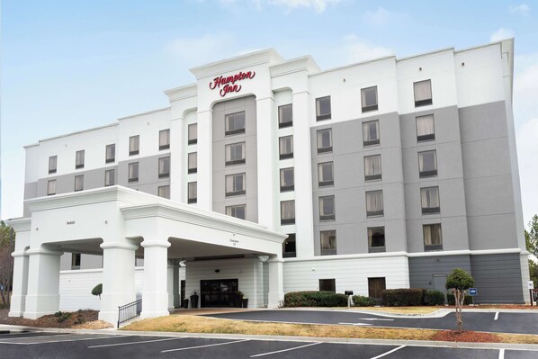 A large, multi-story hotel with a prominent 'Hampton Inn' sign. It features a central entrance with a covered area and multiple windows on each floor.