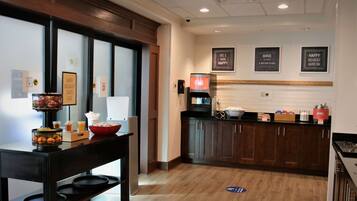 Zona de desayuno de hotel con una mesa de buffet, una cafetera y un refrigerador.