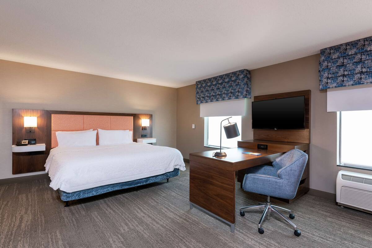 A hotel room with a large bed, a desk with a chair, a flat-screen TV, and a window with blinds.