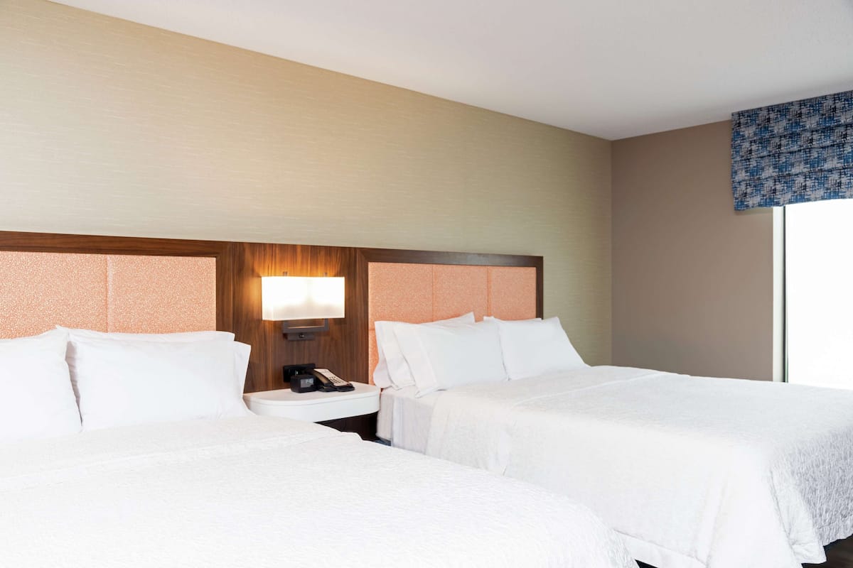 A hotel room with two beds, a nightstand with a lamp, and a window with a blue patterned curtain.