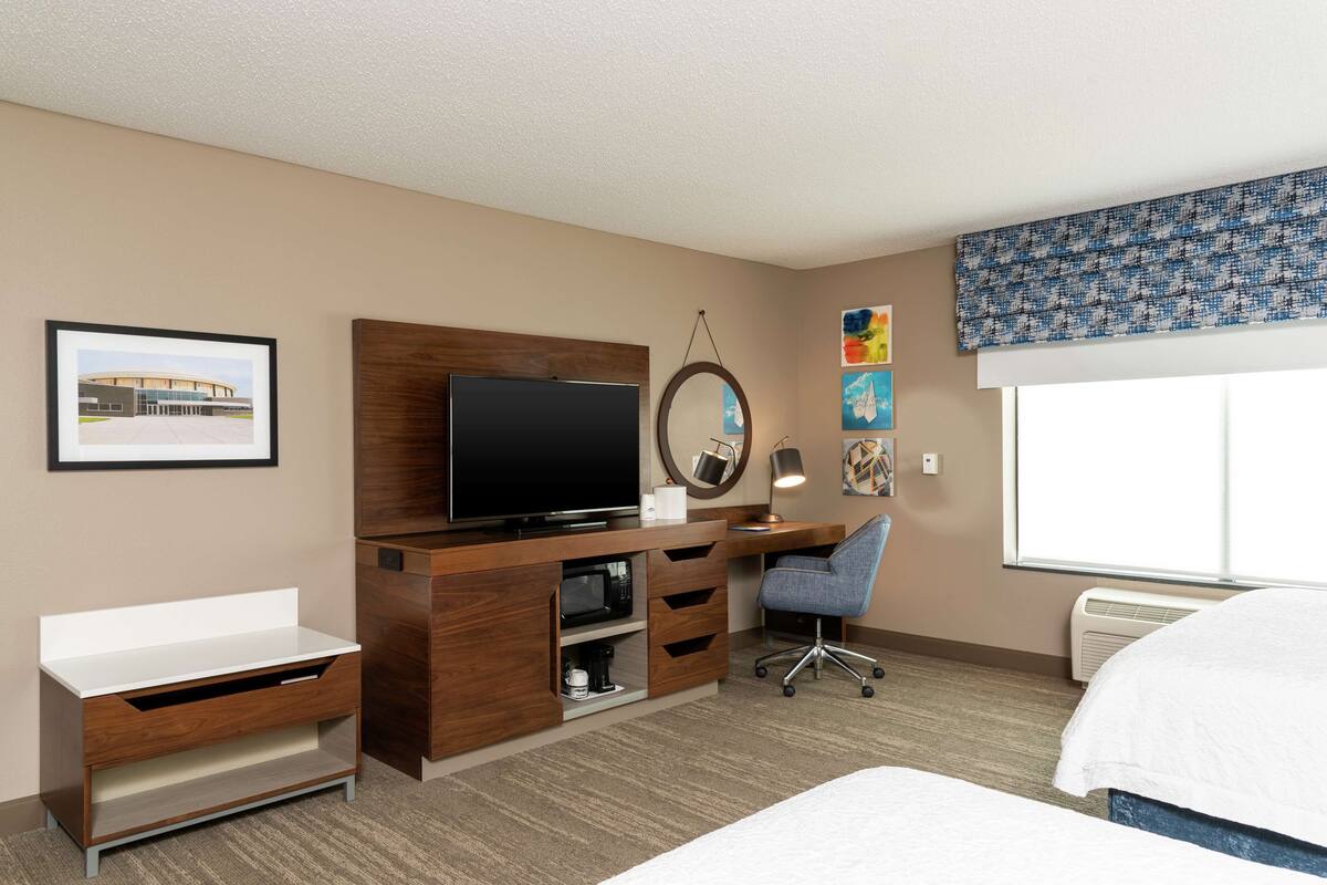 A hotel room with a bed, a desk with a TV, a chair, a microwave, and a large window.