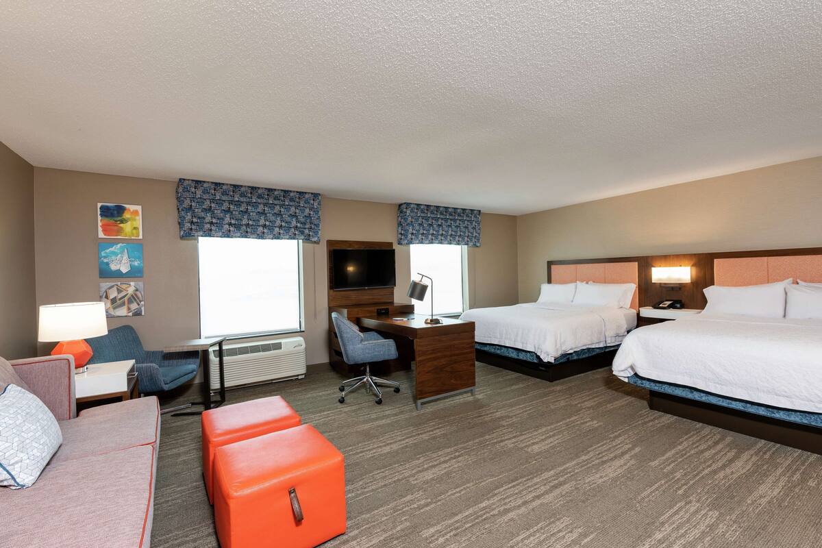 A hotel room with two beds, a desk, a chair, a sofa, and a small orange ottoman.